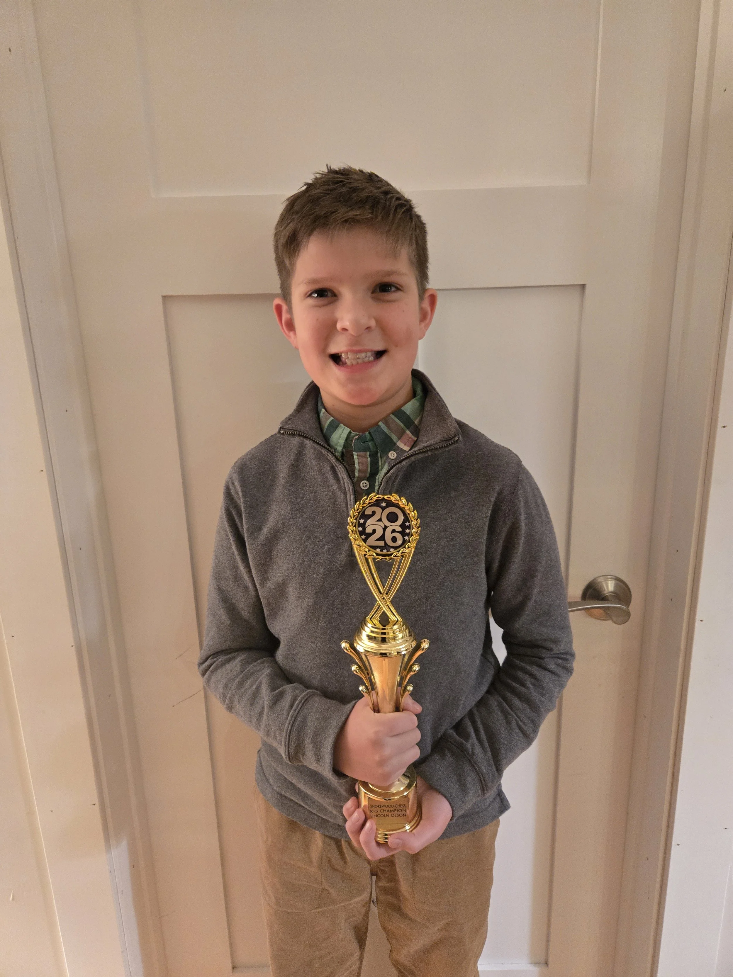 A young boy with short brown hair smiling and holding a gold chess tournament trophy in front of a light-colored door.