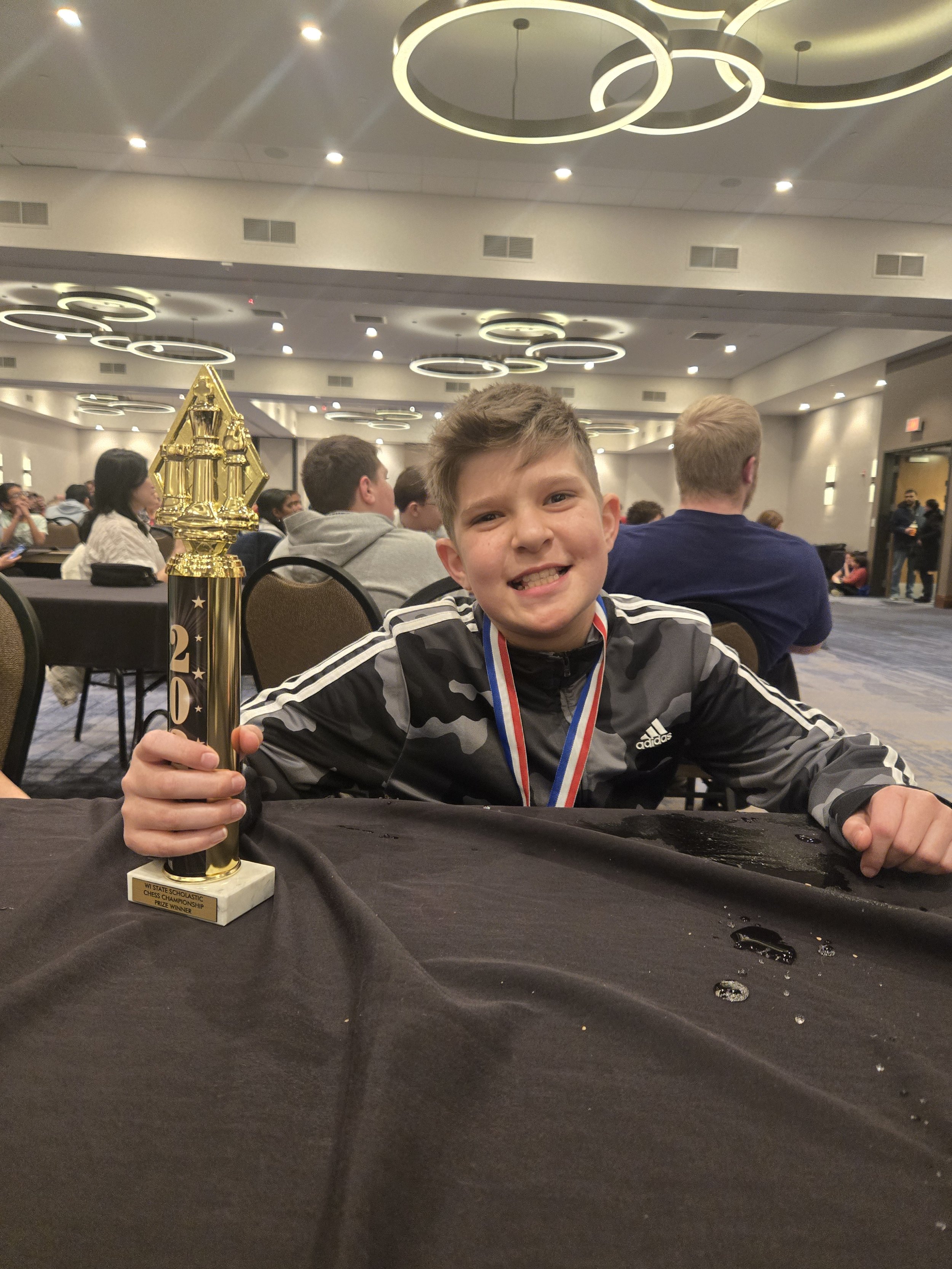 A young boy smiling and holding a trophy at an indoor chess competition. He is wearing a medal and a black and gray sports jacket. The trophy is gold-colored with a chess piece on top and a plaque that reads 'Wisconsin Scholastic Chess Championship Prize Winner.'