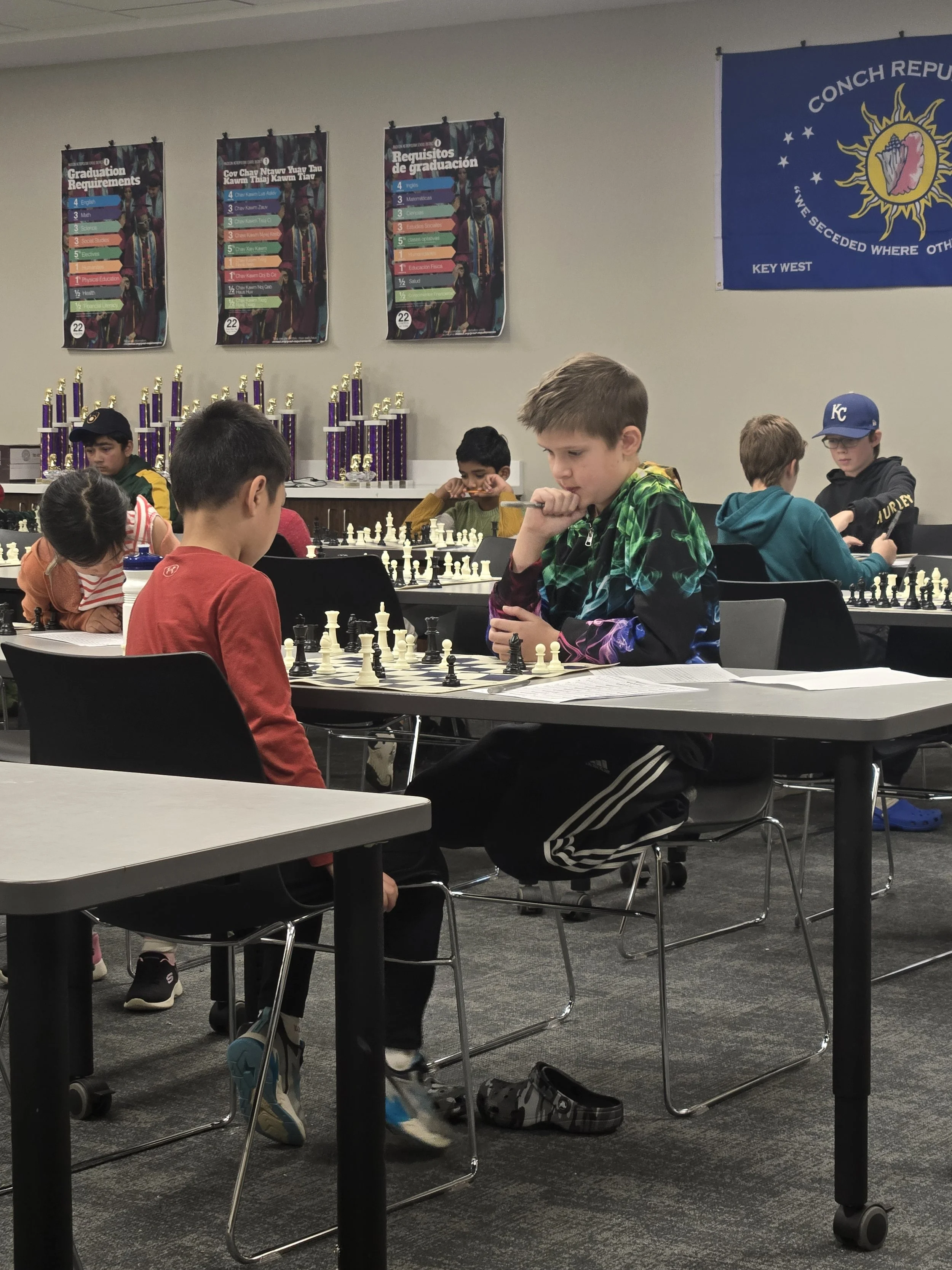 A group of children playing chess indoors, with trophies on shelves behind them, and a blue flag on the wall with a sun and star design.