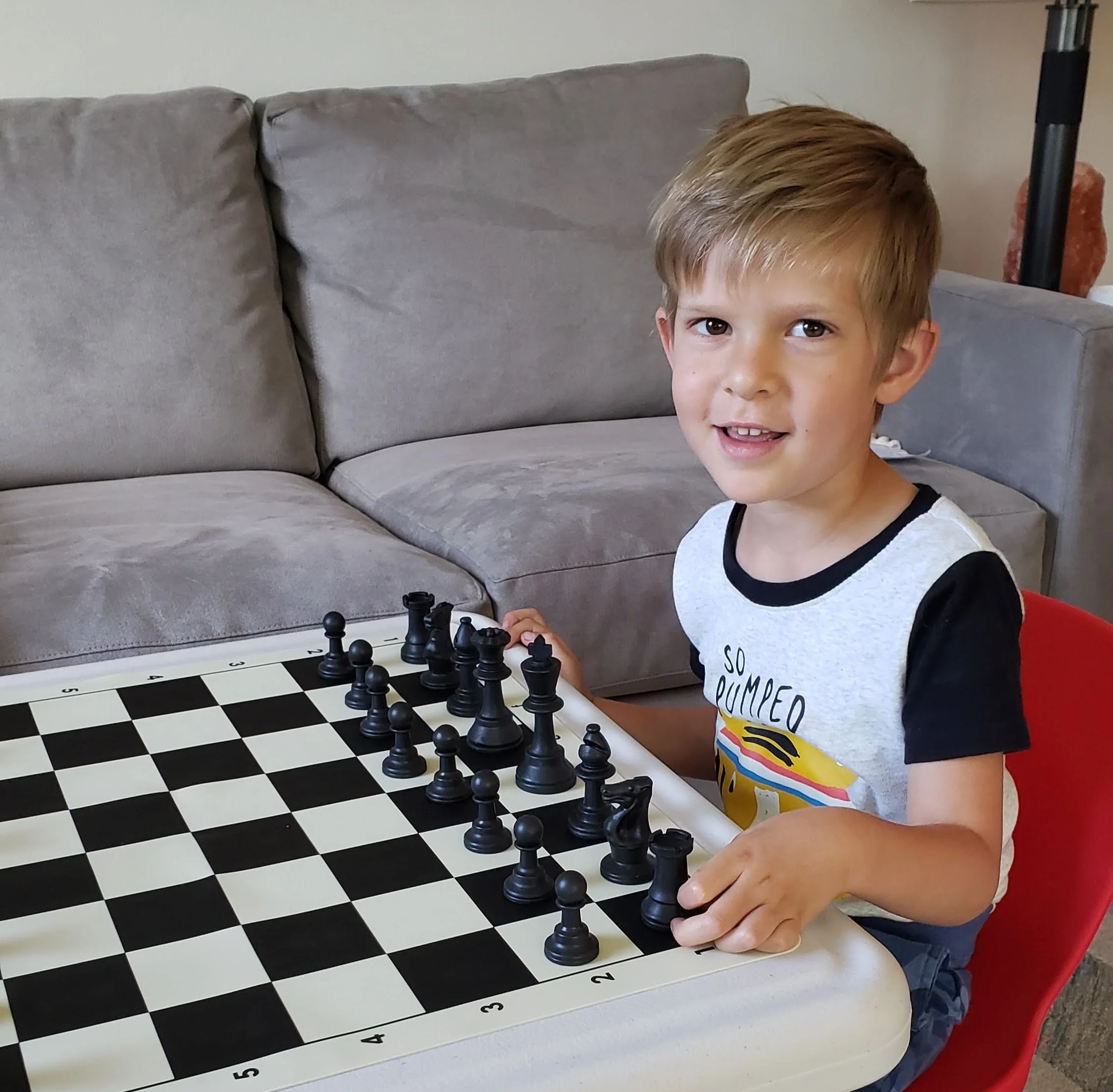 A young boy with light brown hair and a big smile, sitting at a chessboard with a chess game in progress. The chessboard is on a table with black and white squares and numbered rows. The boy is wearing a white and black-shirt with colorful designs on it, and is seated on a red chair. In the background, there is a gray sofa and a salt lamp on a side table.