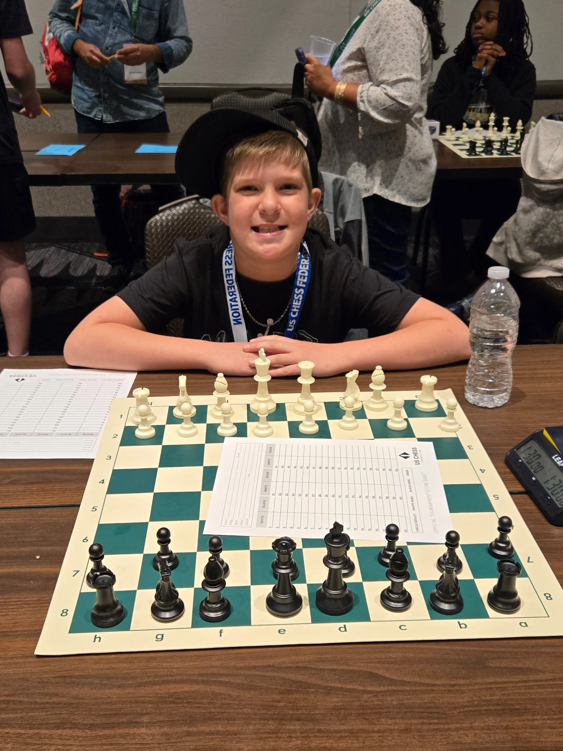 Young boy sitting at a table with a chessboard set up for a game, smiling at the camera. Other people are in the background, some standing and talking, near a table with a chess clock and chess pieces.