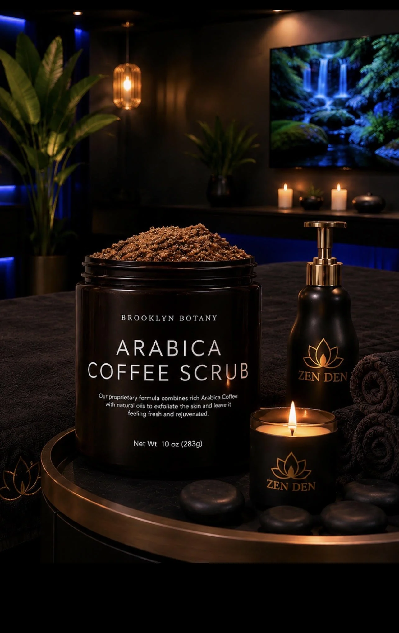 A dark, cozy spa or relaxation room featuring a large container of Arabica coffee scrub labeled Brooklyn Botany, a gold pump bottle labeled Zen Den, a small lit candle, candles in the background, green plants, a large TV screen displaying a waterfall scene, and a round tray with black stones.