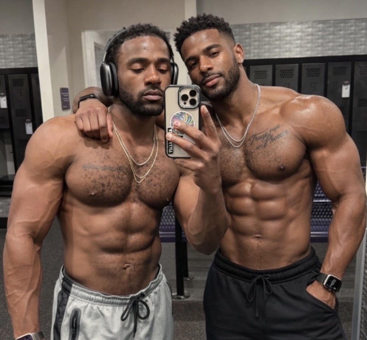 Two muscular shirtless men with tattoos taking a selfie in a gym locker room, one wearing headphones, the other with a smartwatch, both flexing their abs and looking at the camera.