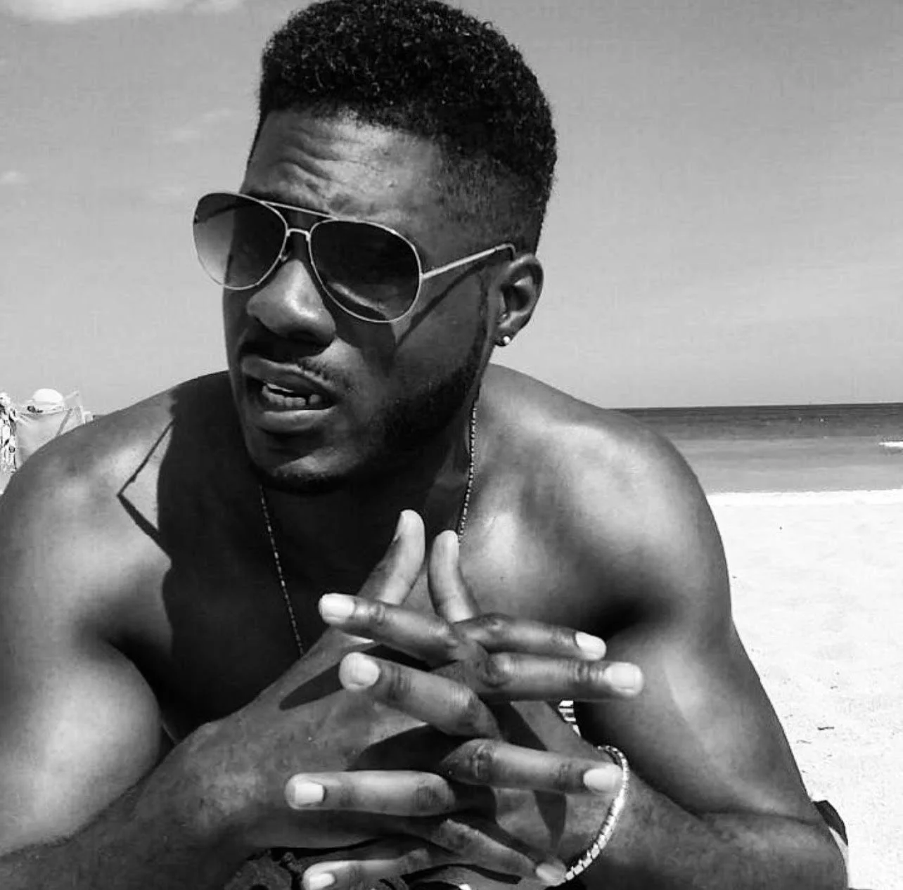 A shirtless young man wearing sunglasses and jewelry is at the beach, with the ocean and sandy shore in the background.