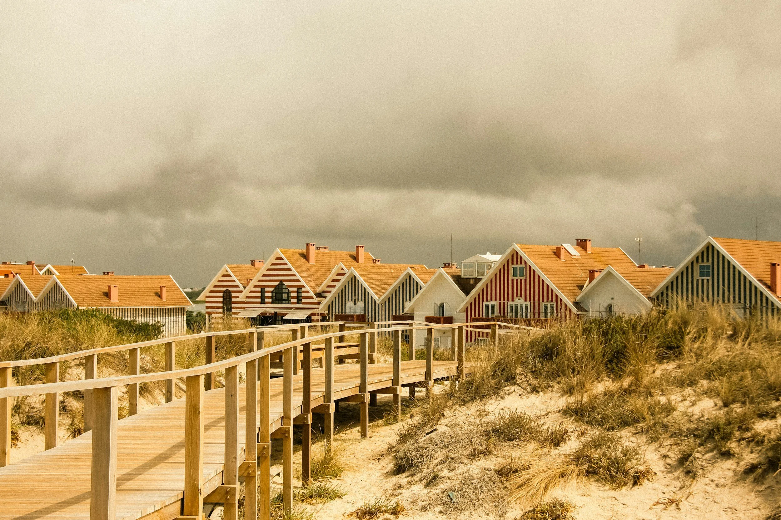 Row of colorful beach houses with striped facades, a wooden boardwalk, and dune grass under a cloudy sky.