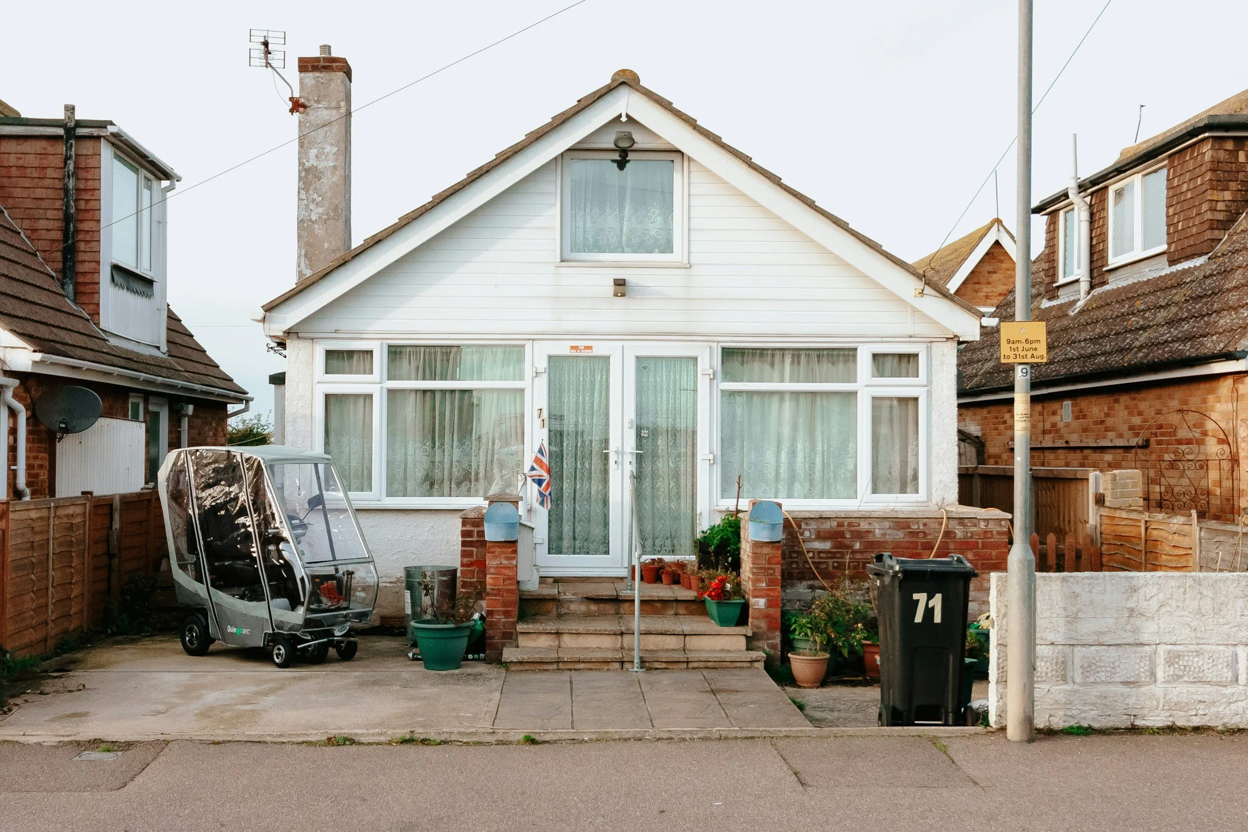 Front view of a white house with large windows and a small roof attic window, surrounded by other brick houses, with a driveway and various household items in the yard, including a baby stroller, potted plants, and a black garbage bin.