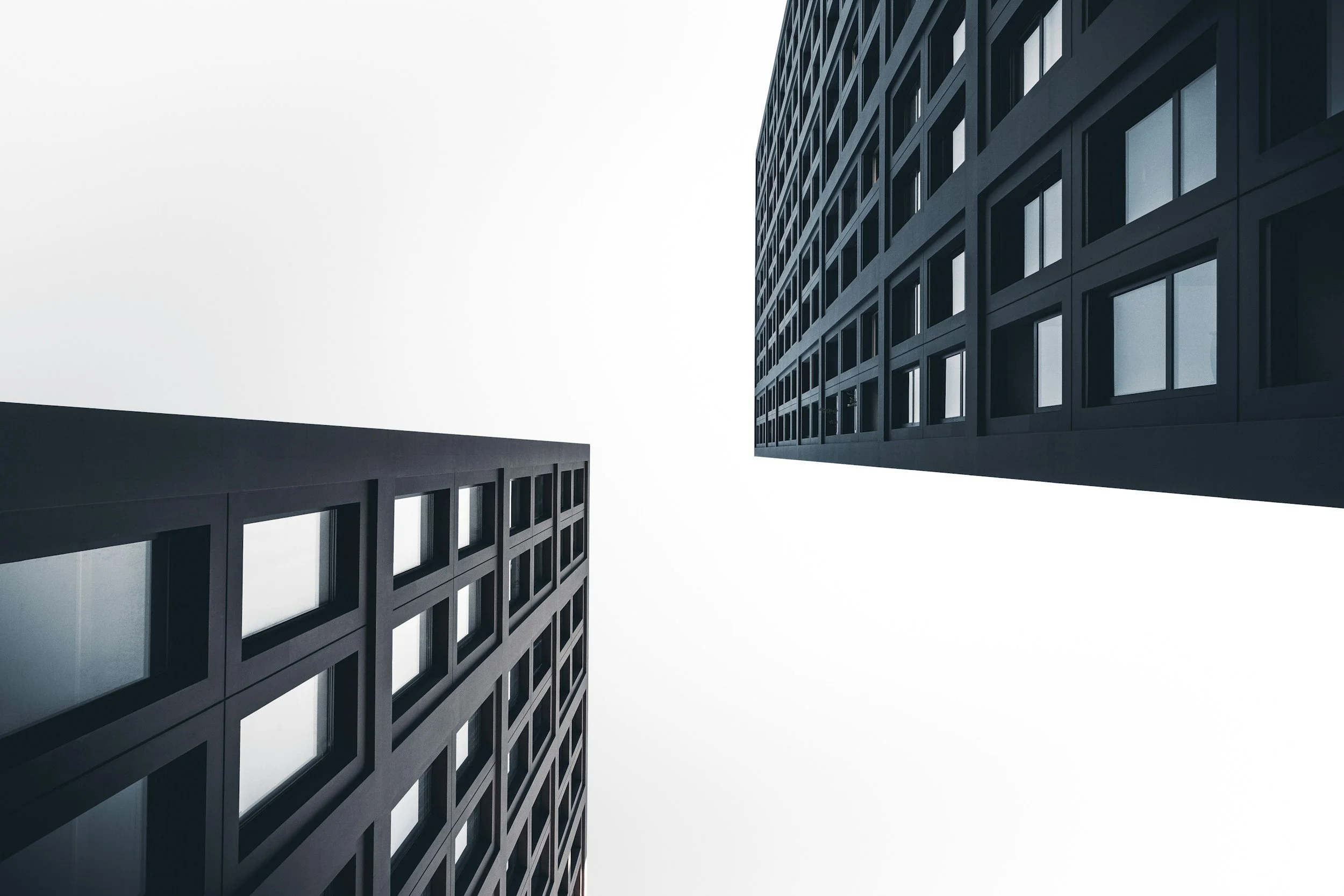 Low-angle view of two modern office buildings with grid-like window patterns against an overcast sky.