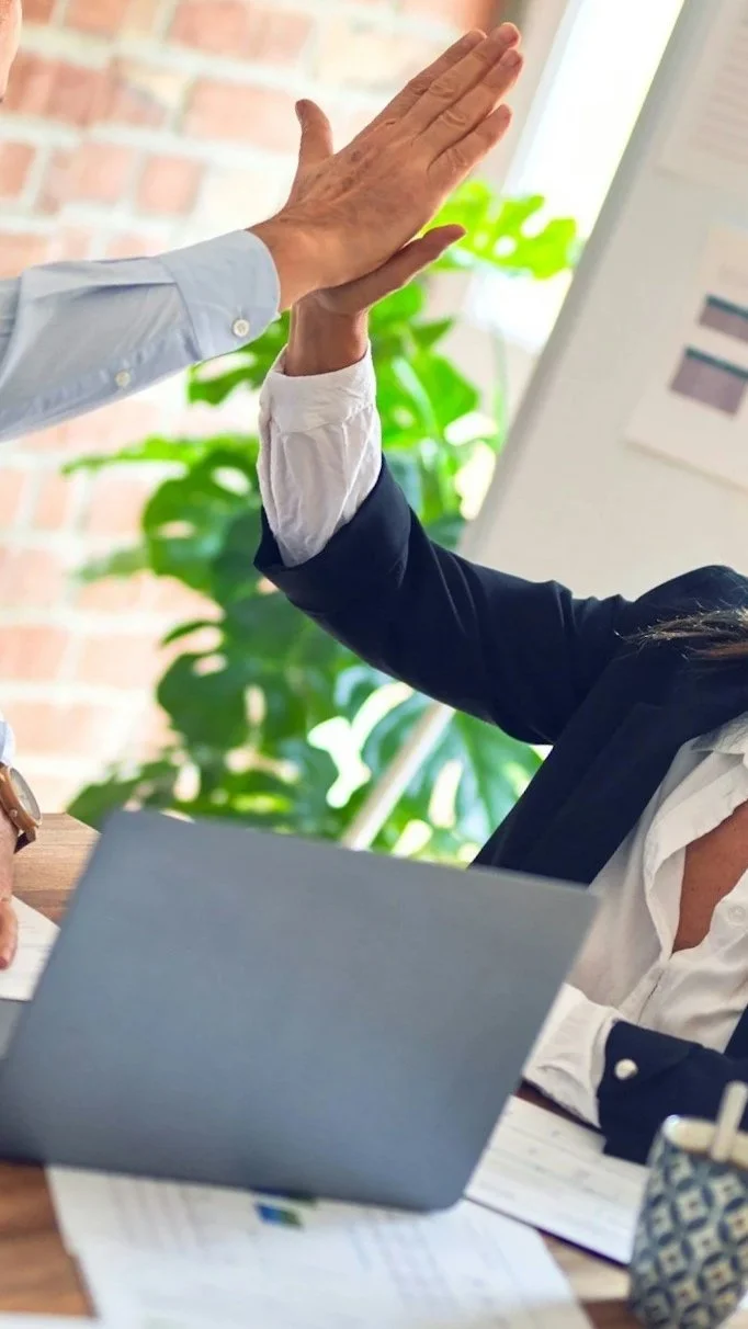 Two people giving each other a high five in an office setting with a laptop, papers, and a plant in the background.
