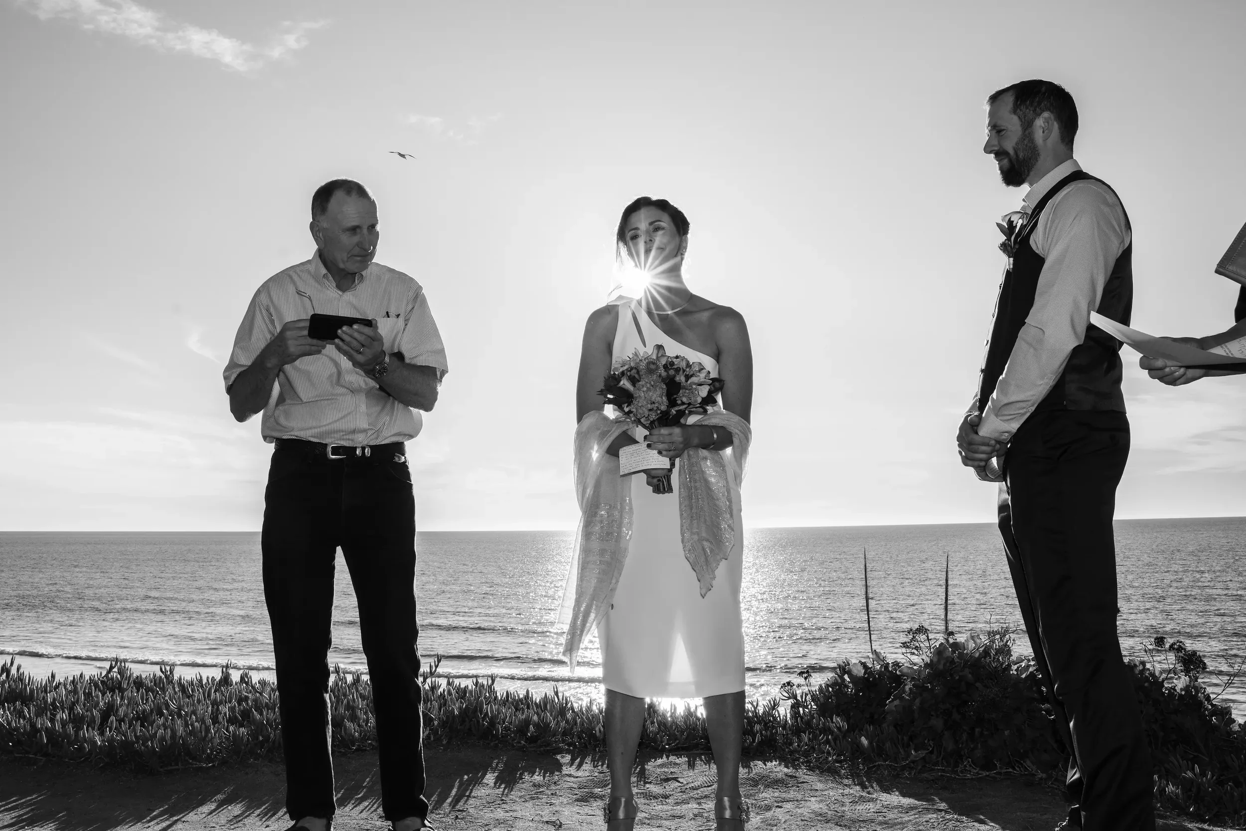 Black and white photo of a wedding ceremony at the beach, with three people: a bride holding a bouquet, a groom, and a officiant, with the ocean and sky in the background.