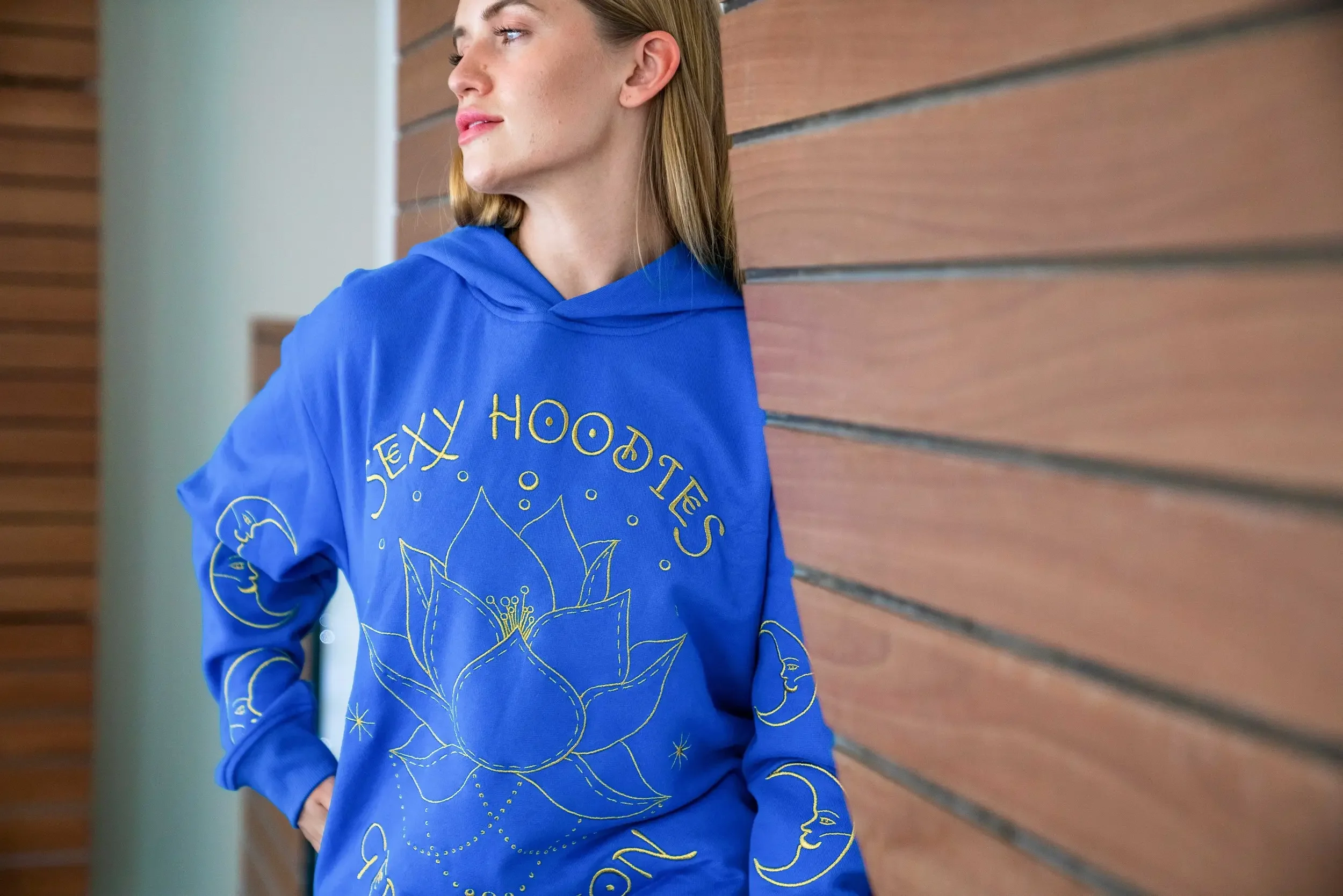 A woman in a blue hoodie with yellow line art design leaning against a wooden wall, looking to her left with hands in her pockets.