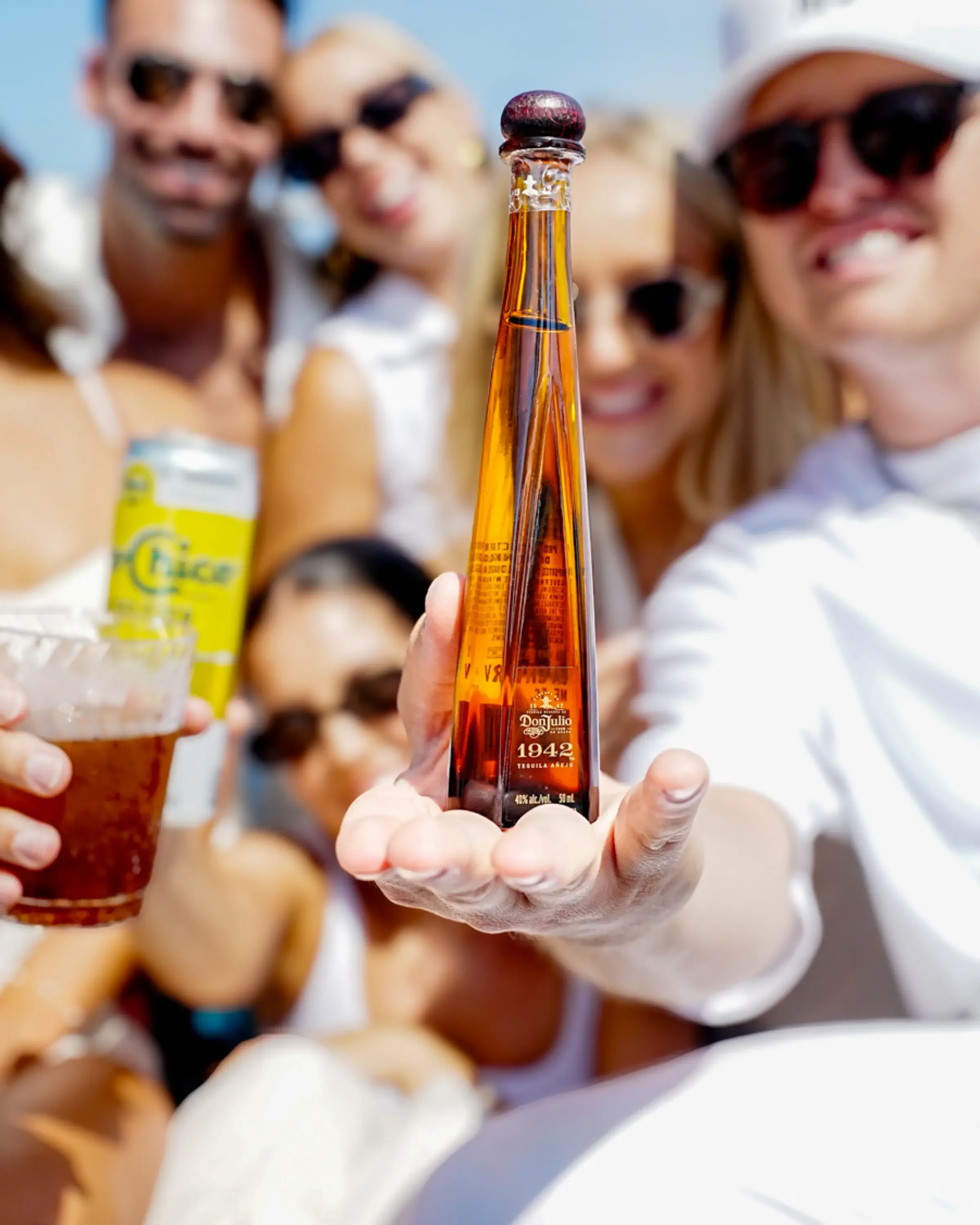 Close-up of a hand holding a bottle of Don Julio tequila in front of a group of smiling women wearing sunglasses at an outdoor gathering.