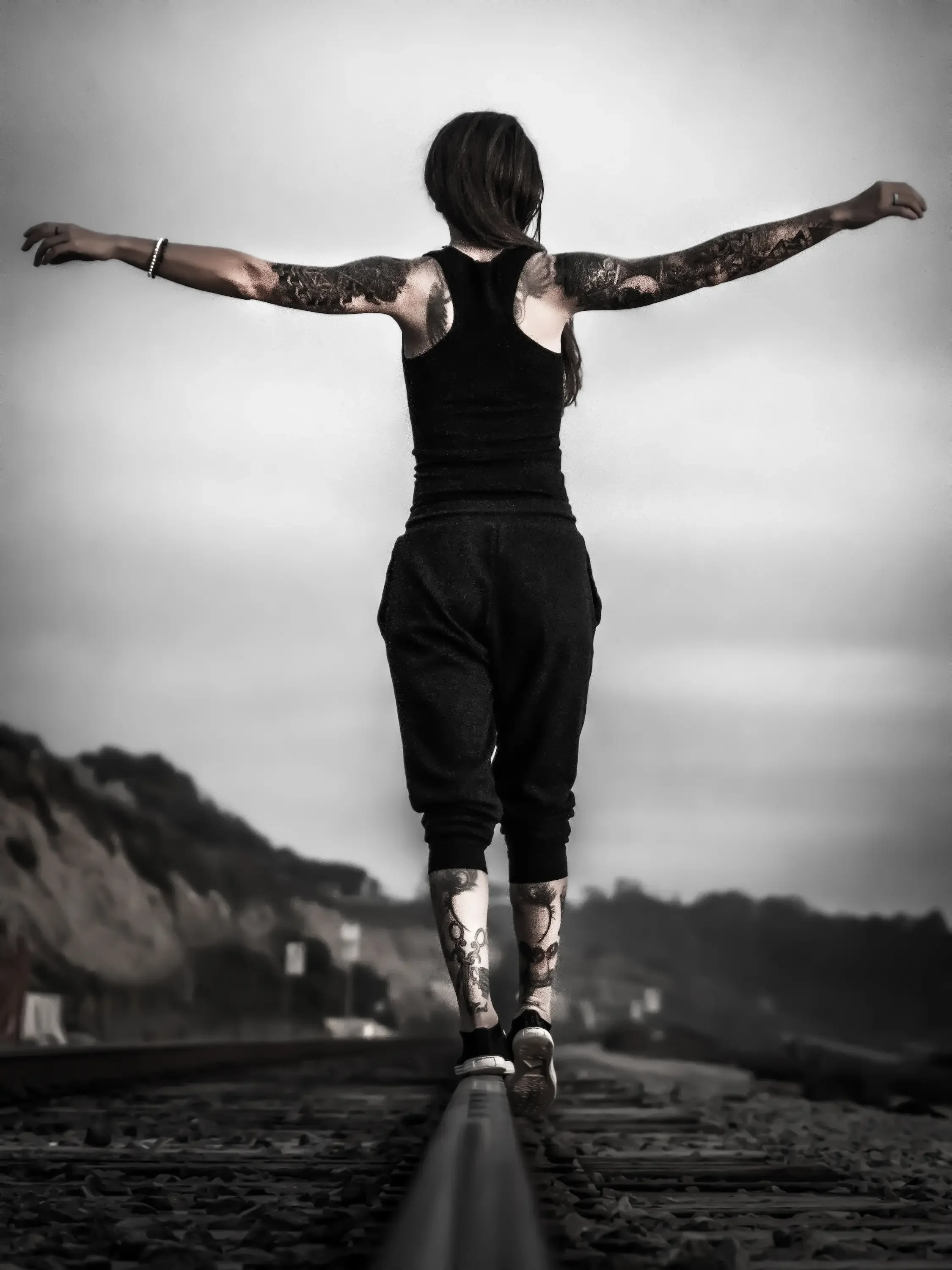 A person with tattoos on their arms and legs, dressed in black, balancing on a railway track with arms outstretched, facing away from the camera, against a cloudy sky background.