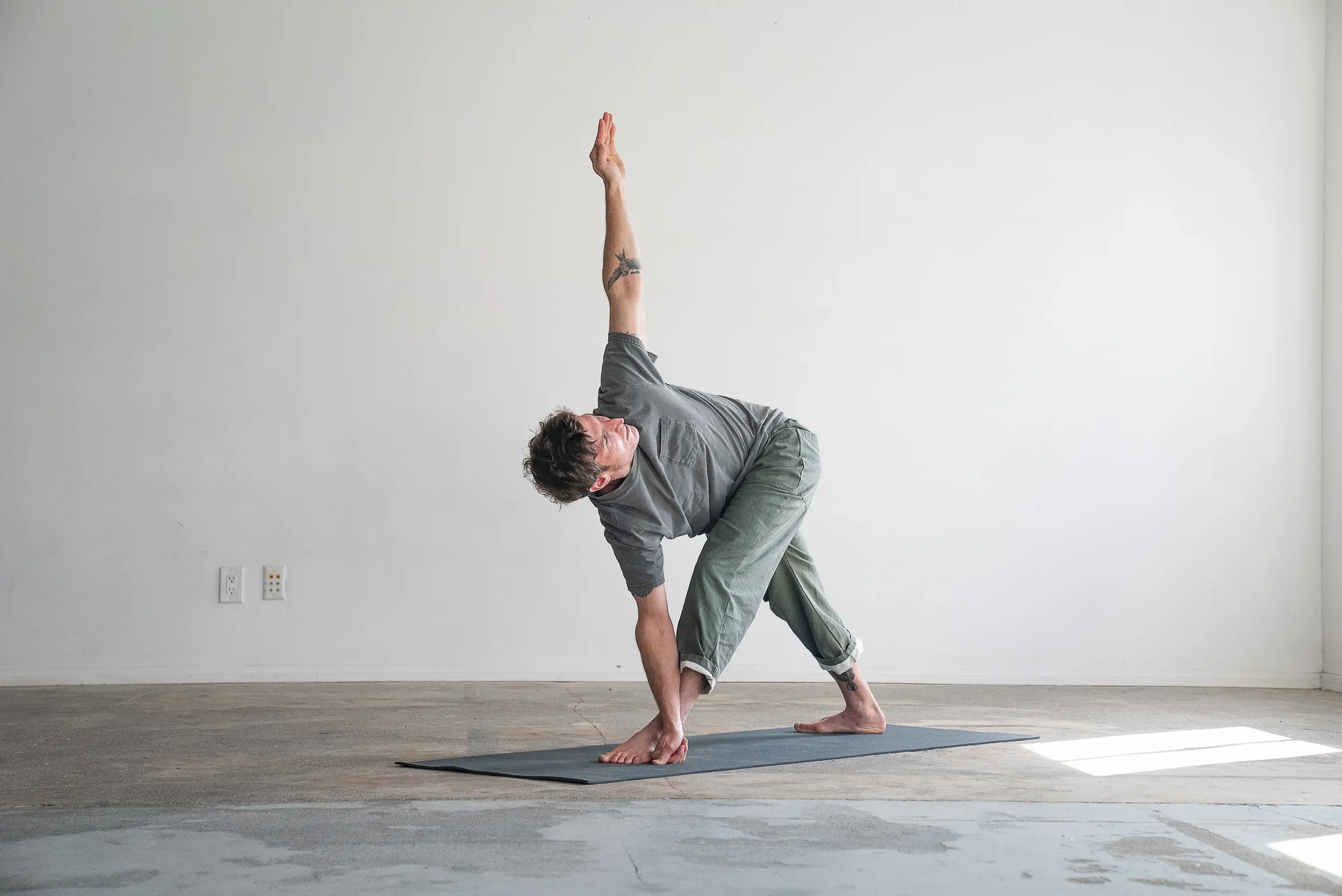 A man with tattoos practicing yoga in a studio, holding a balance pose with one hand on the floor and one arm raised, on a exercise mat.
