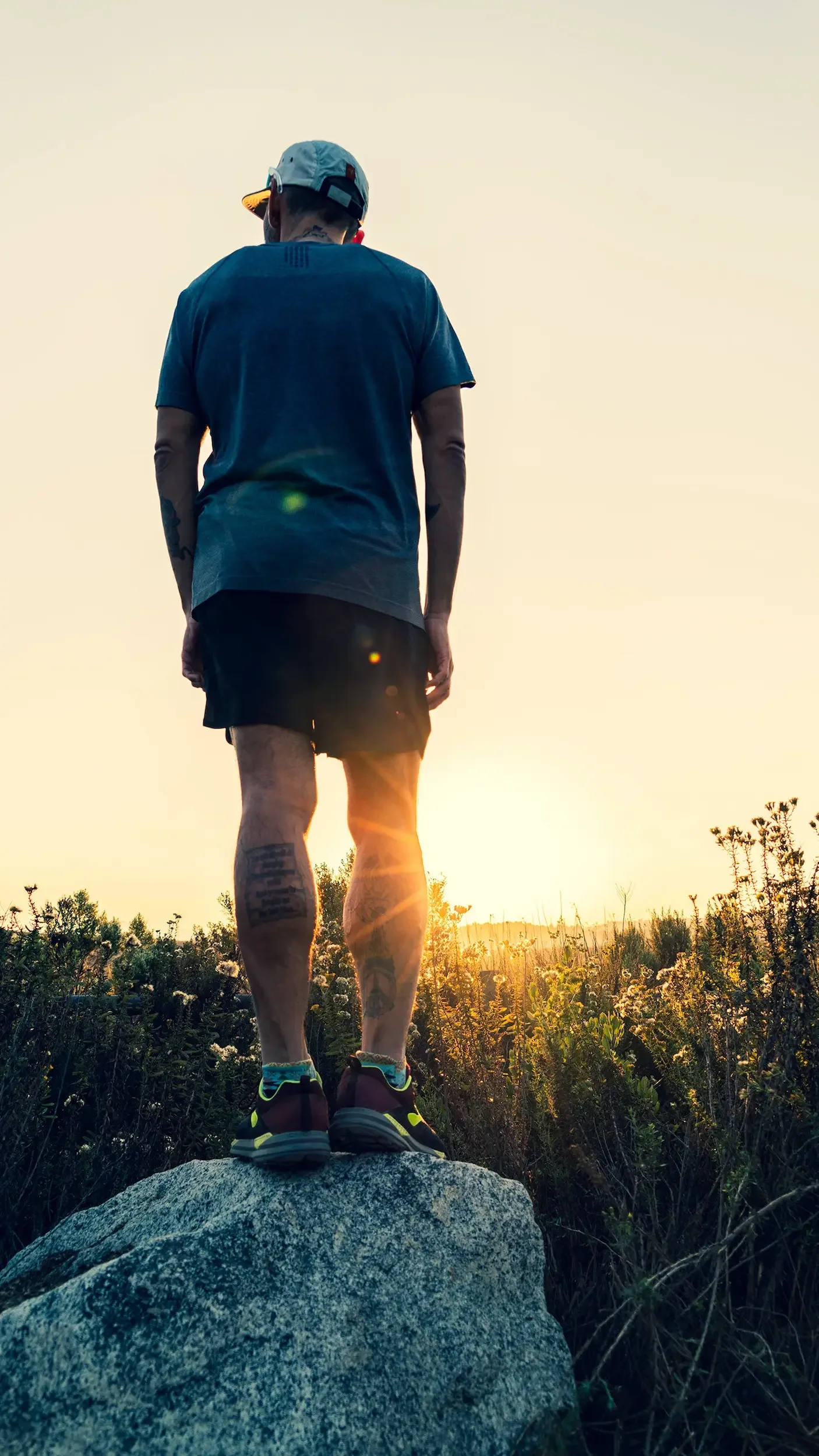 A man standing on a large rock outdoors during sunset, wearing a cap, t-shirt, shorts, and sneakers, watching the horizon with a field of plants and trees around him.