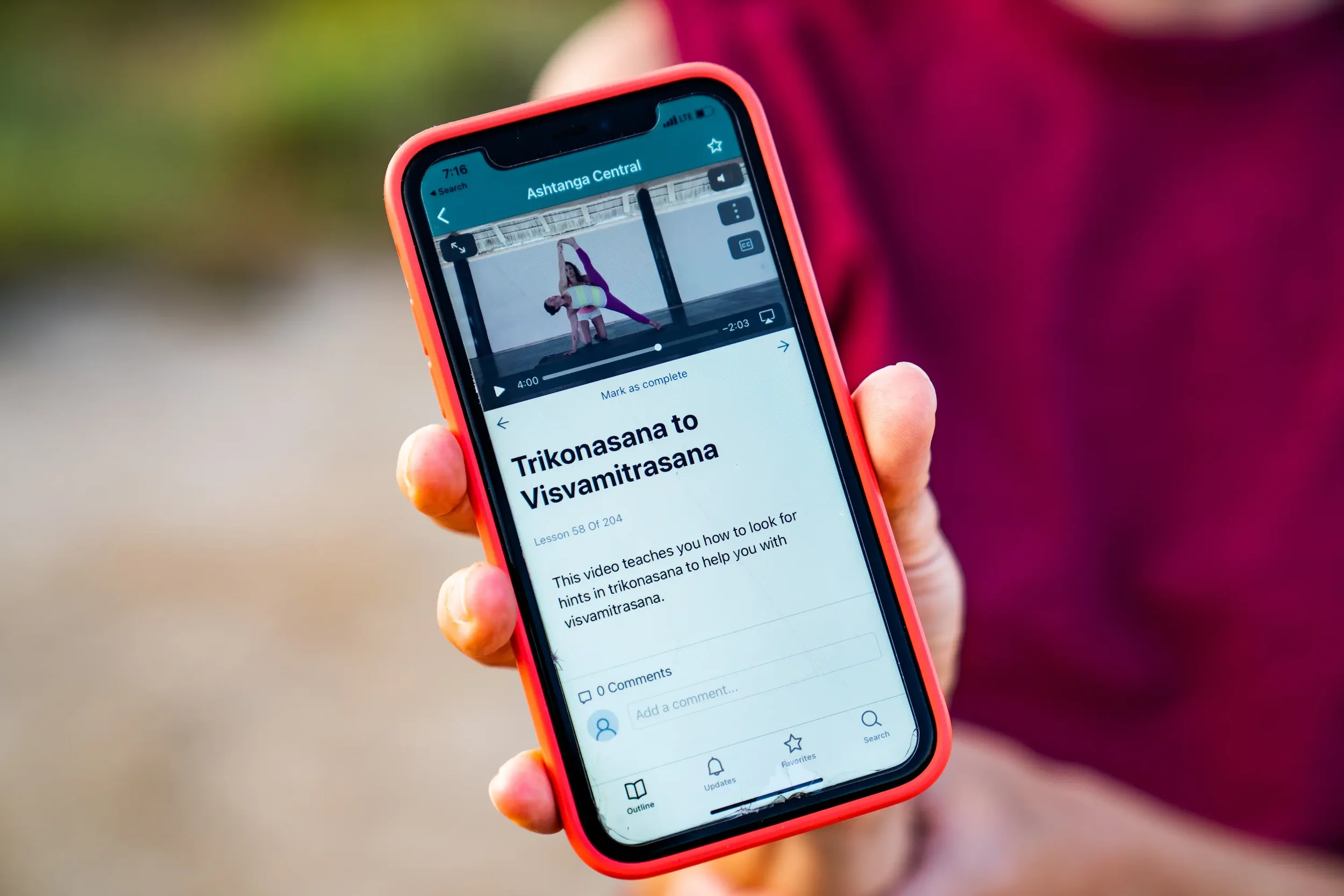 Close-up of a person's hand holding a smartphone in pink case, displaying an online yoga class titled 'Trikonasana to Visvamitrasana' on the screen, with a video of two women practicing yoga in a studio.