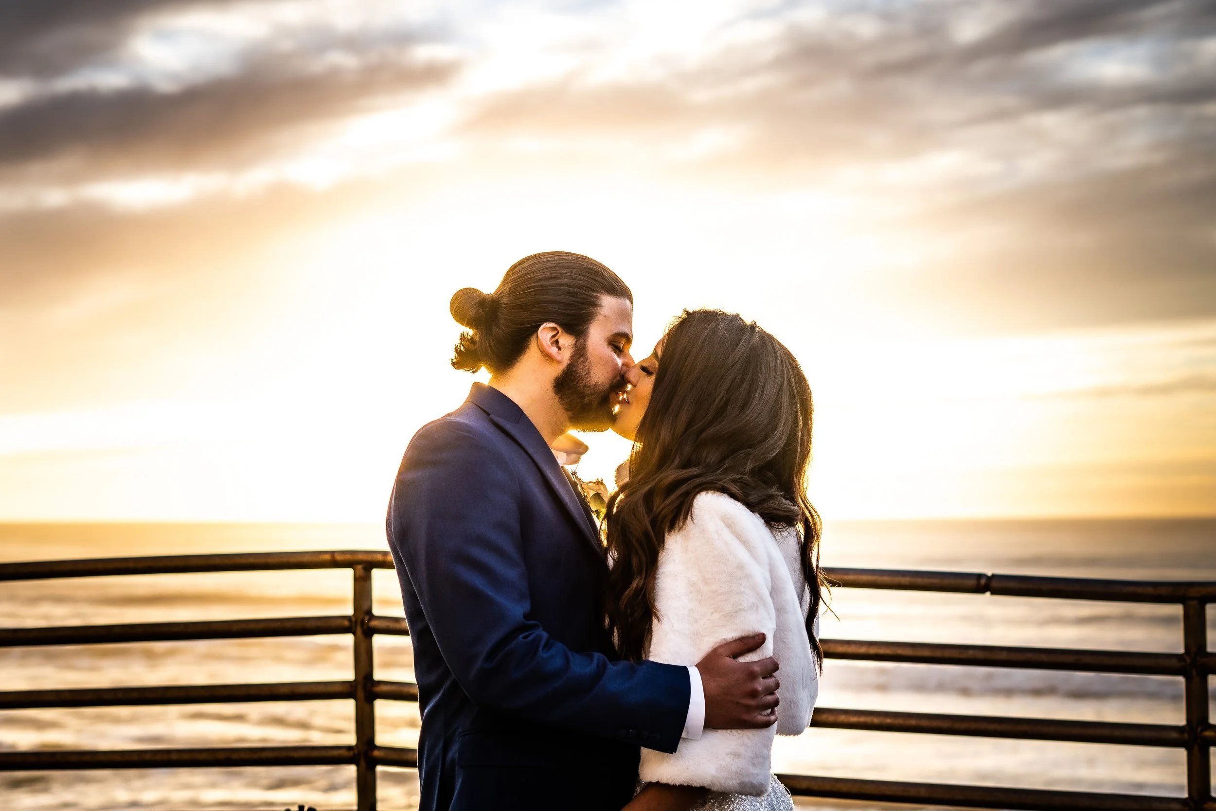 A couple is kissing on a pier by the water during sunset, with a cloudy sky in the background.