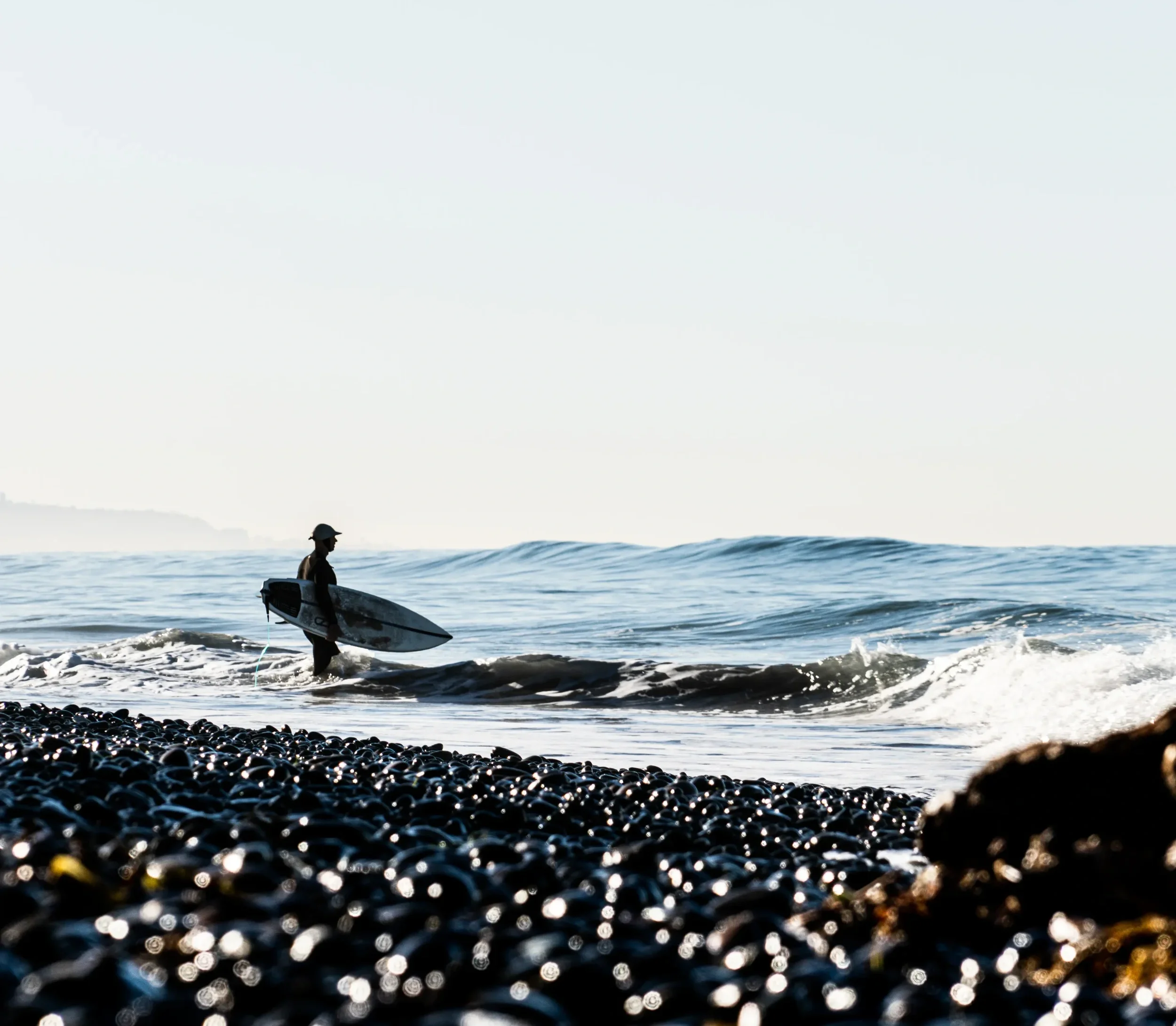 Person carrying surfboard walking on rocky shoreline towards ocean waves during sunset.