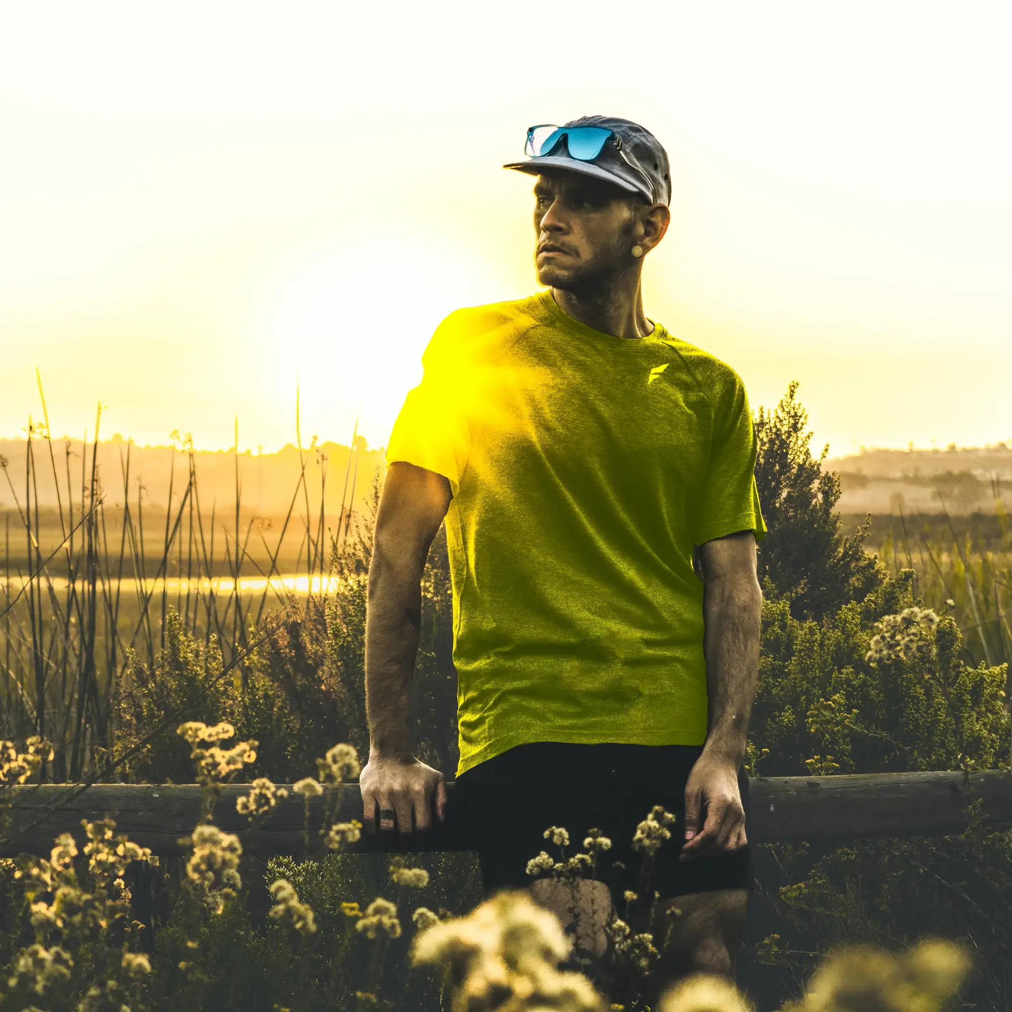 A man wearing a gray cap with sunglasses on top, a yellow shirt, and black shorts stands outdoors during sunset or sunrise, surrounded by flowers and greenery.