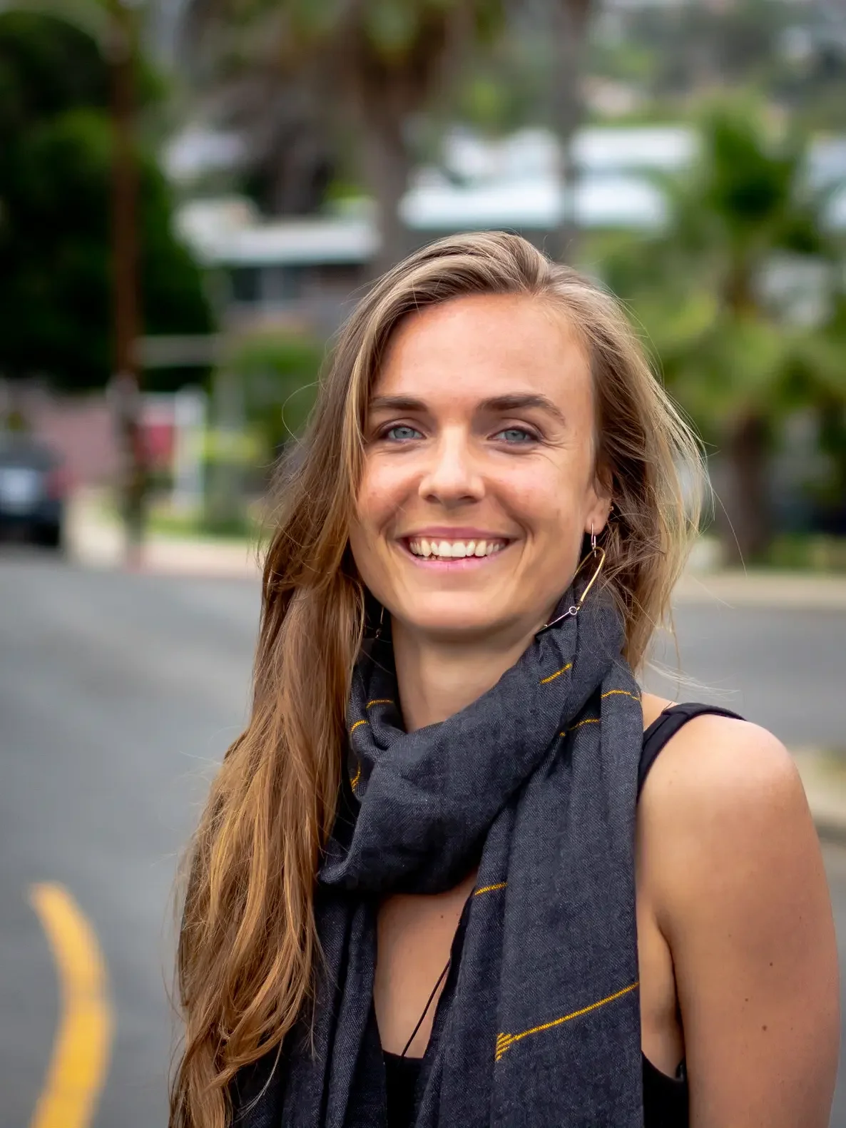 A smiling woman with long, wavy blonde hair, wearing a black sleeveless top and a dark gray scarf with yellow stripes, standing outdoors on a street with trees and houses in the background.