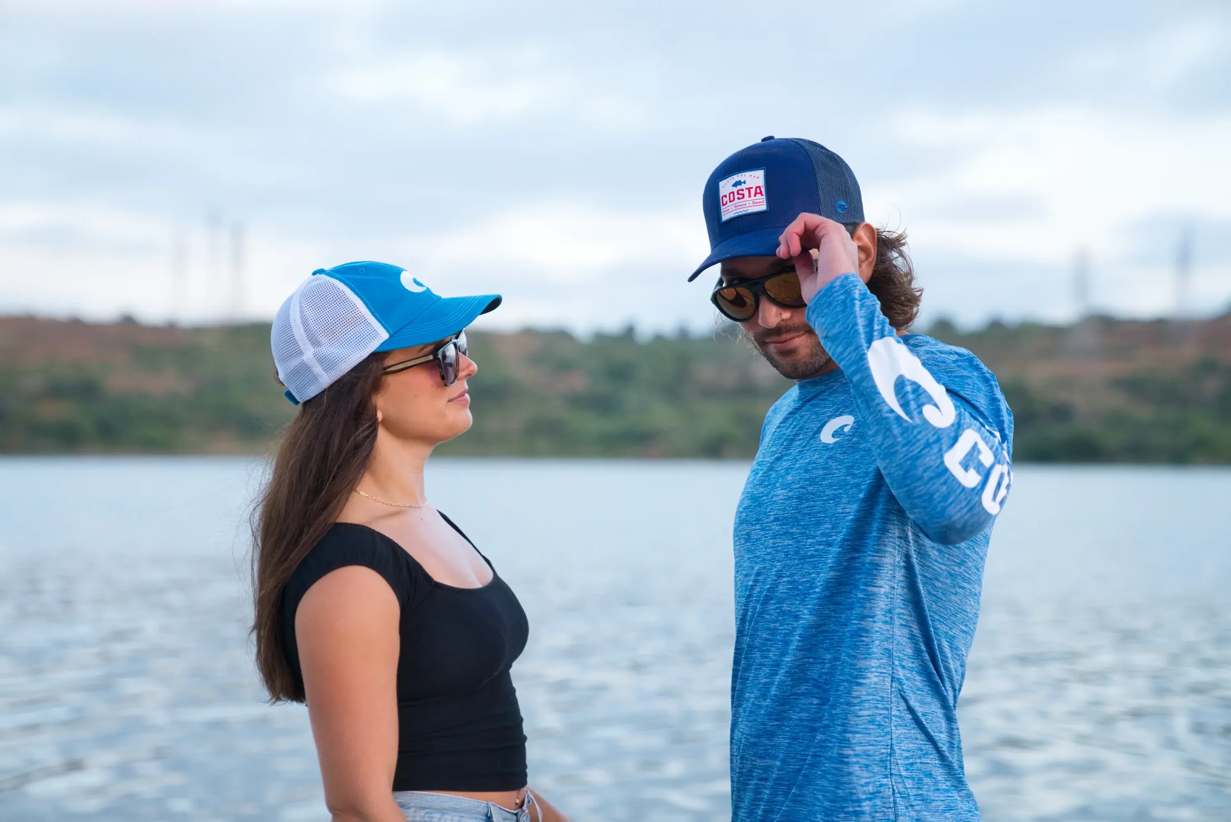 A man and woman standing near a body of water, facing each other, with the man adjusting his cap. The woman is wearing sunglasses and a black top, while the man is wearing sunglasses, a blue cap with a Costa logo, and a blue shirt with white text.