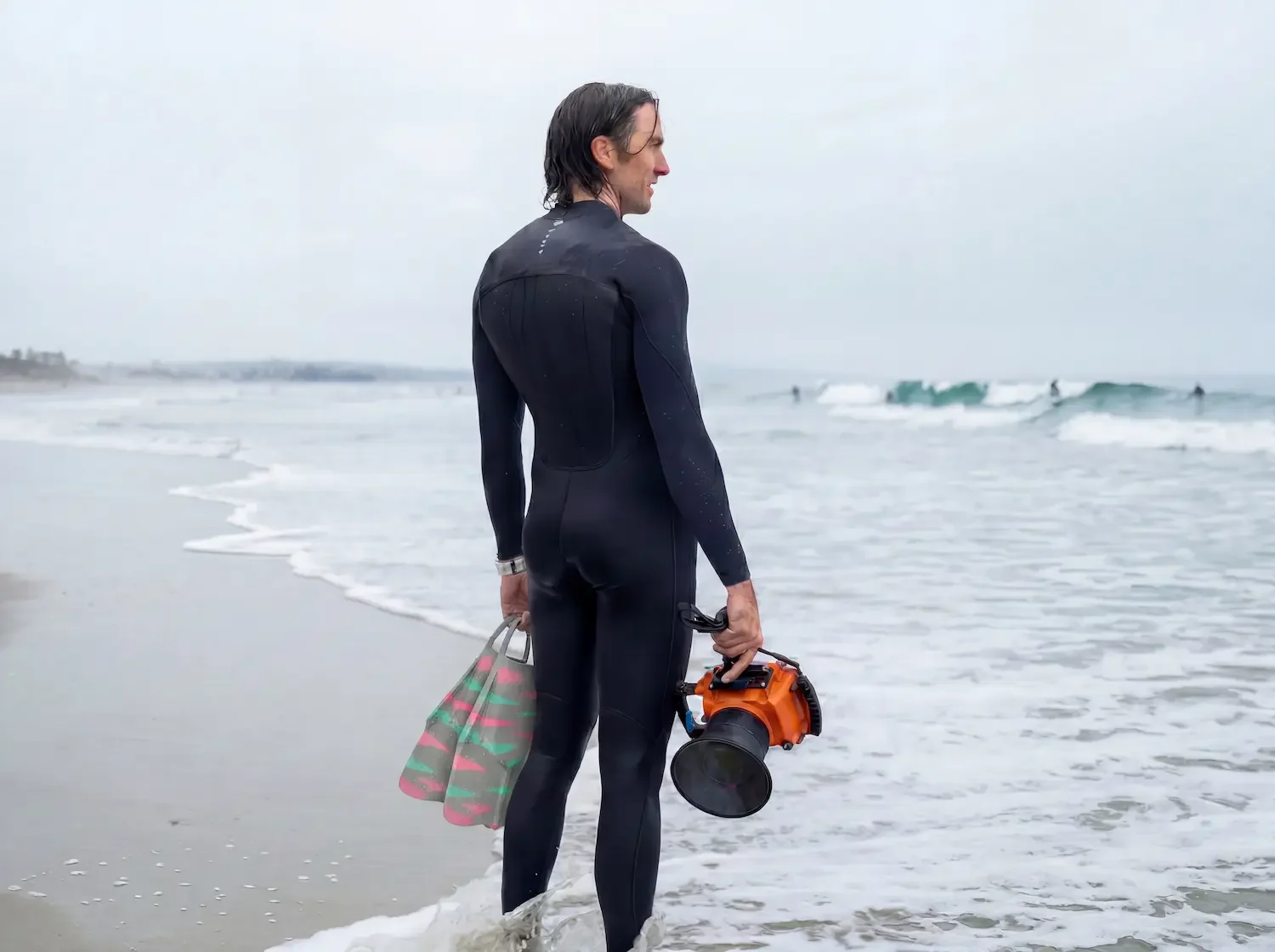 Photographer in a wetsuit carrying swim fins and an underwater camera housing, walking toward the surf