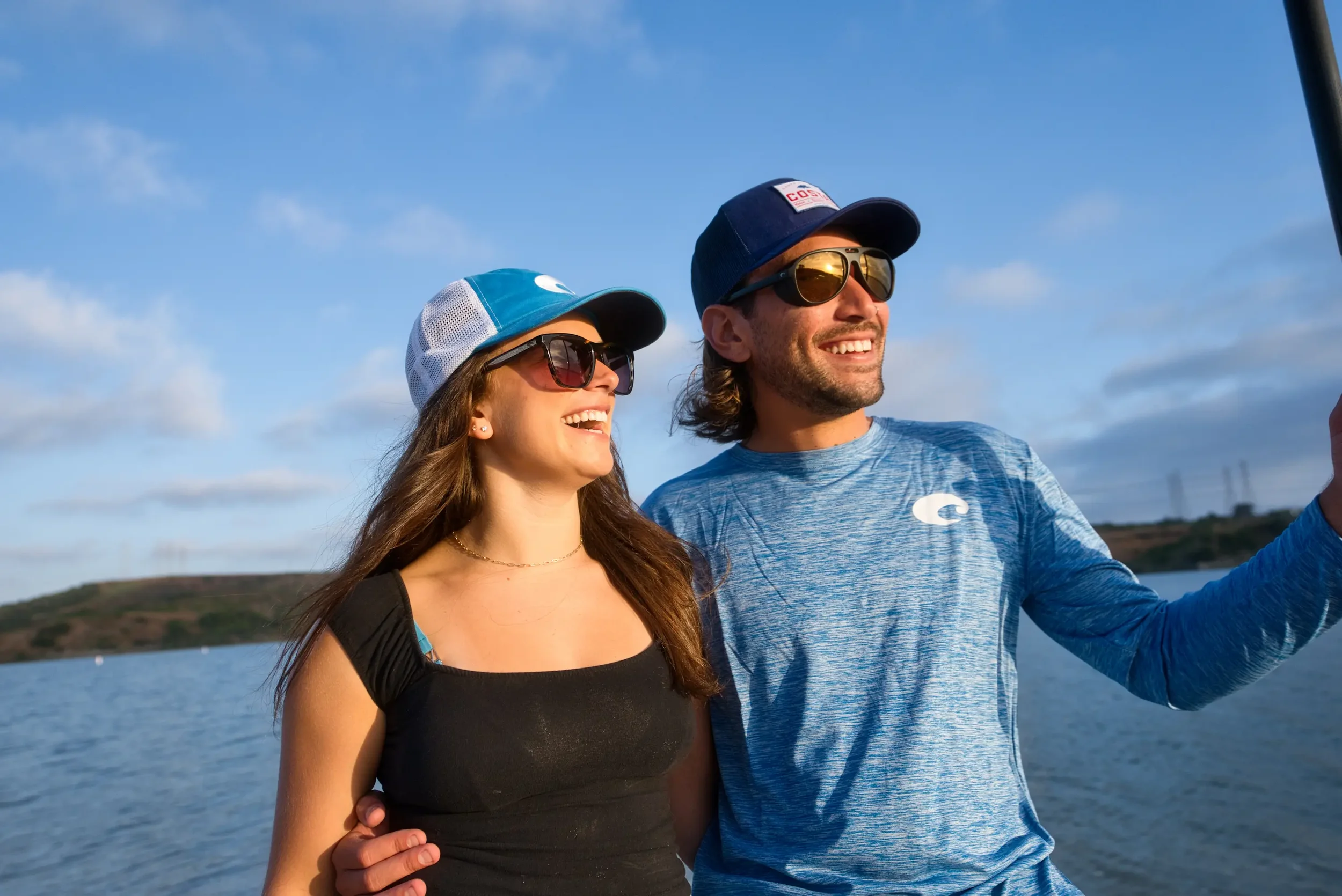 A young woman and man, both wearing sunglasses and hats, enjoying a boat ride on a lake or river under a partly cloudy sky, with a forested shoreline in the background.
