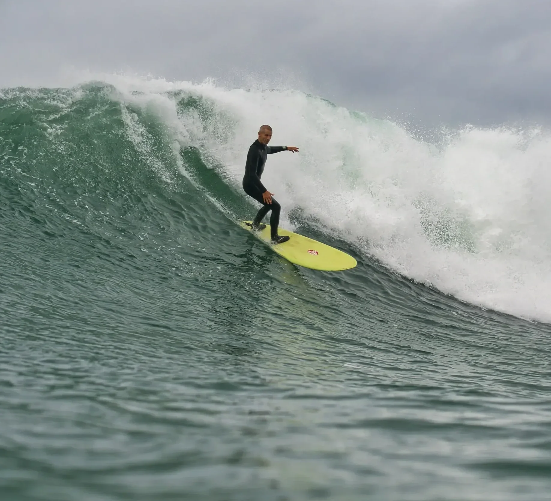 A man in a wetsuit surfing on a yellow surfboard on a large ocean wave under an overcast sky.