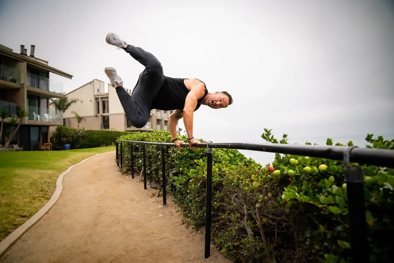 Man performing a one-arm handstand on a railing outdoors, with greenery and residential buildings in the background.