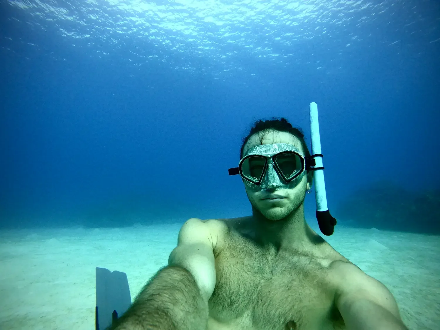 A man taking a selfie underwater wearing goggles and a snorkel, with coral and ocean water in the background.