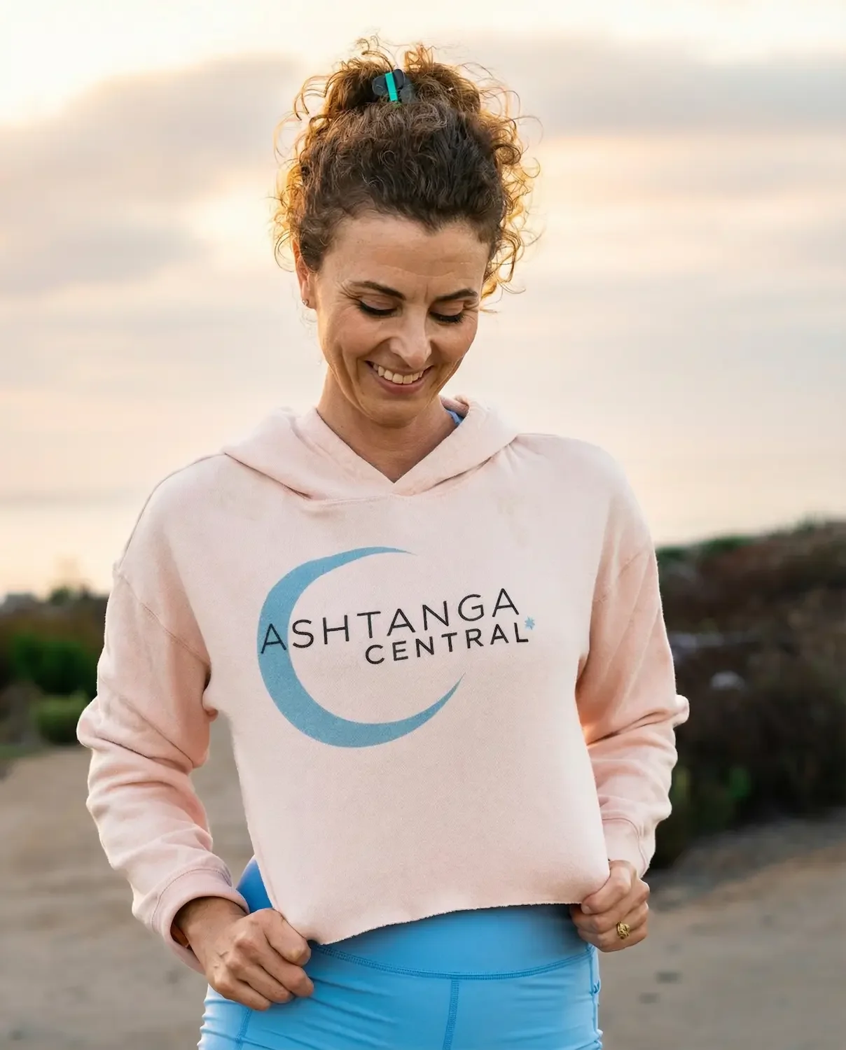 Woman smiling outdoors wearing a pink hoodie with a moon and Stars logo and the words Ashtanga Central.