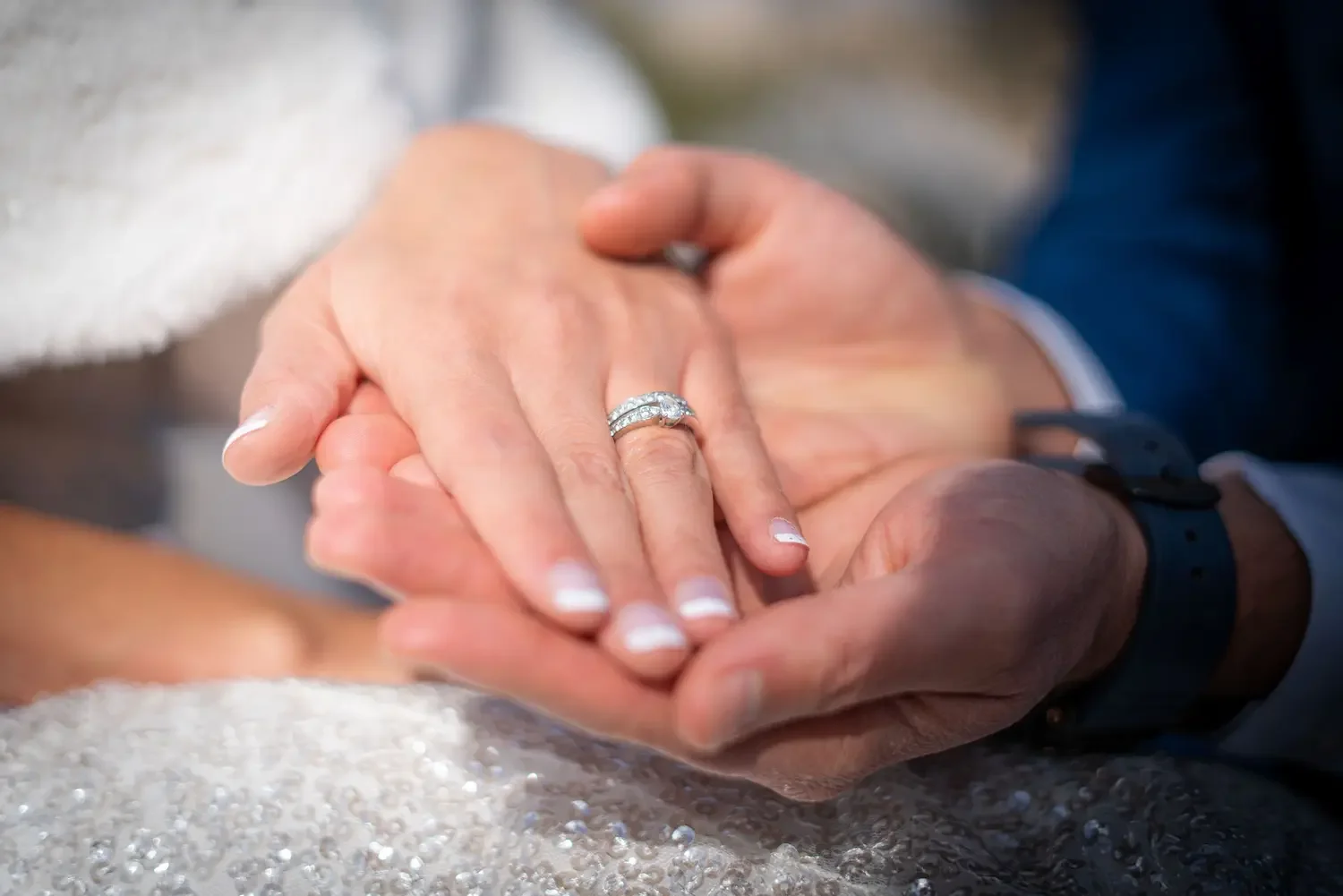 Close-up of a man and woman holding hands, the woman wearing a diamond engagement ring and wedding band, with a blurred background.
