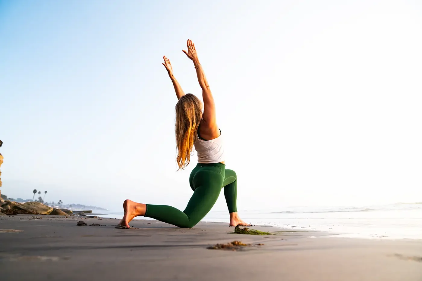 A woman practicing yoga on the beach, performing a lunge pose with arms raised towards the sky during sunrise or sunset.