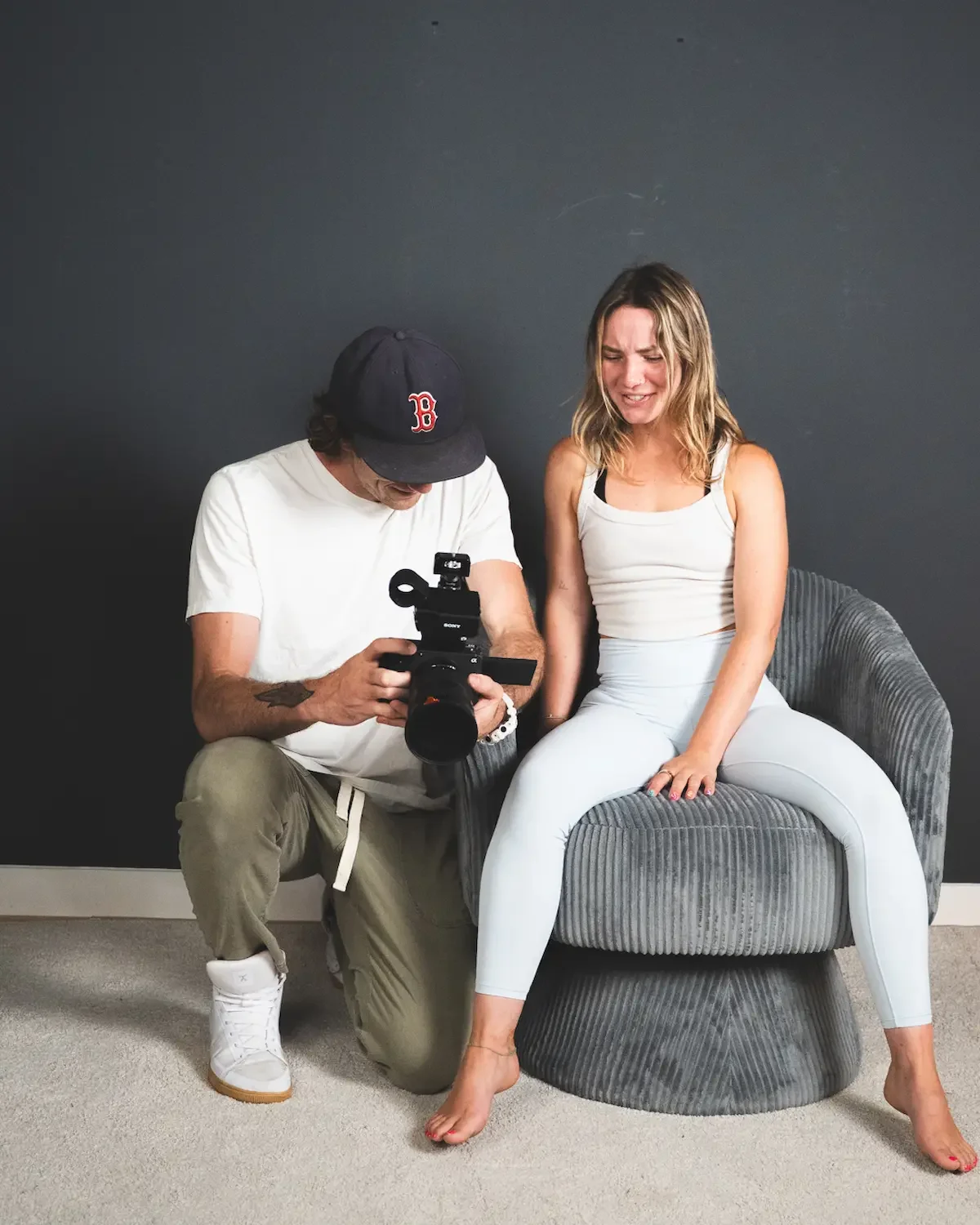 A woman sitting on a gray, rounded armchair with a distressed facial expression, while a man films or photographs her with a professional camera. The woman is wearing a white tank top and white leggings, with barefooted. The man is wearing a white t-shirt, beige pants, white sneakers, and a cap with a red 'B' logo, is kneeling on one knee beside her, holding the camera to his eye.