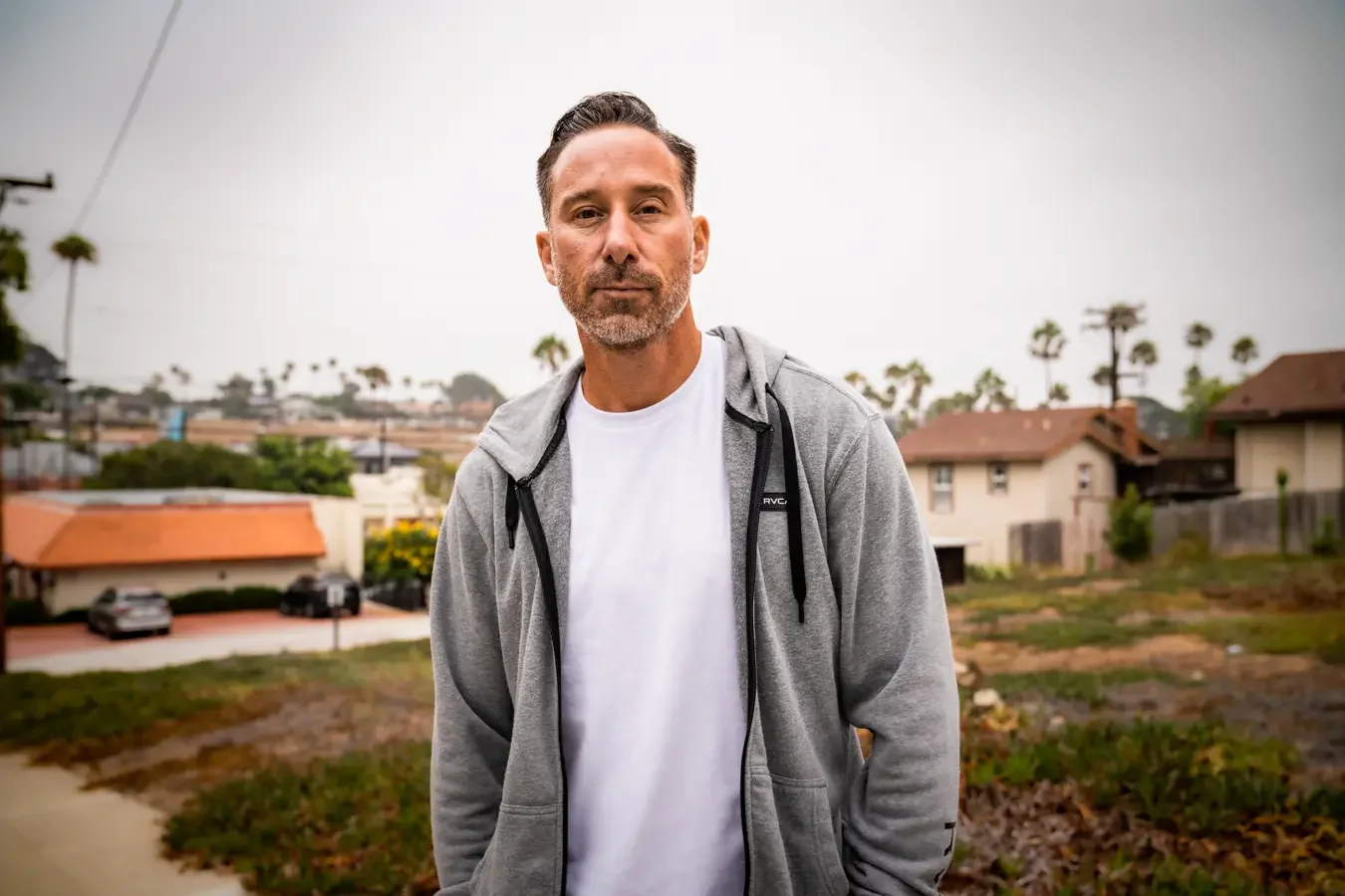 A middle-aged man with a beard in a white t-shirt and gray hoodie standing outdoors with houses, trees, and a cloudy sky in the background.