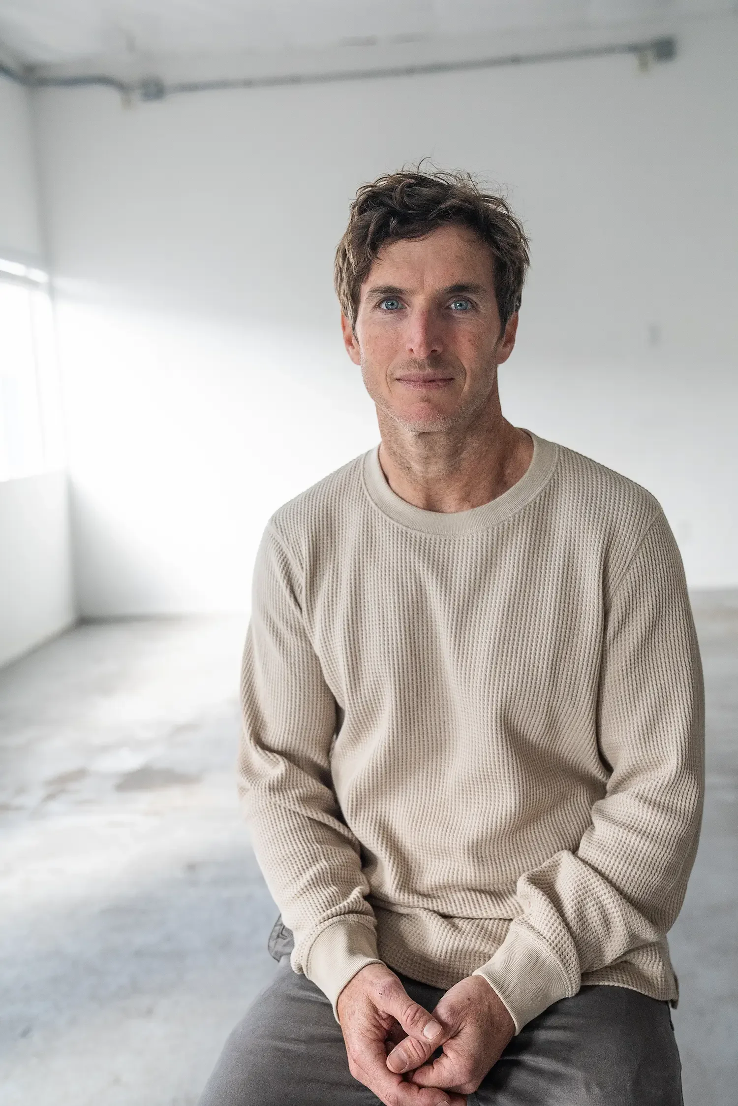 A man with tousled dark hair, light skin, and blue eyes, dressed in a cream-colored sweater and gray pants, sitting on a stool in a bright, minimalist room with white walls and floor, looking directly at the camera with a neutral expression.