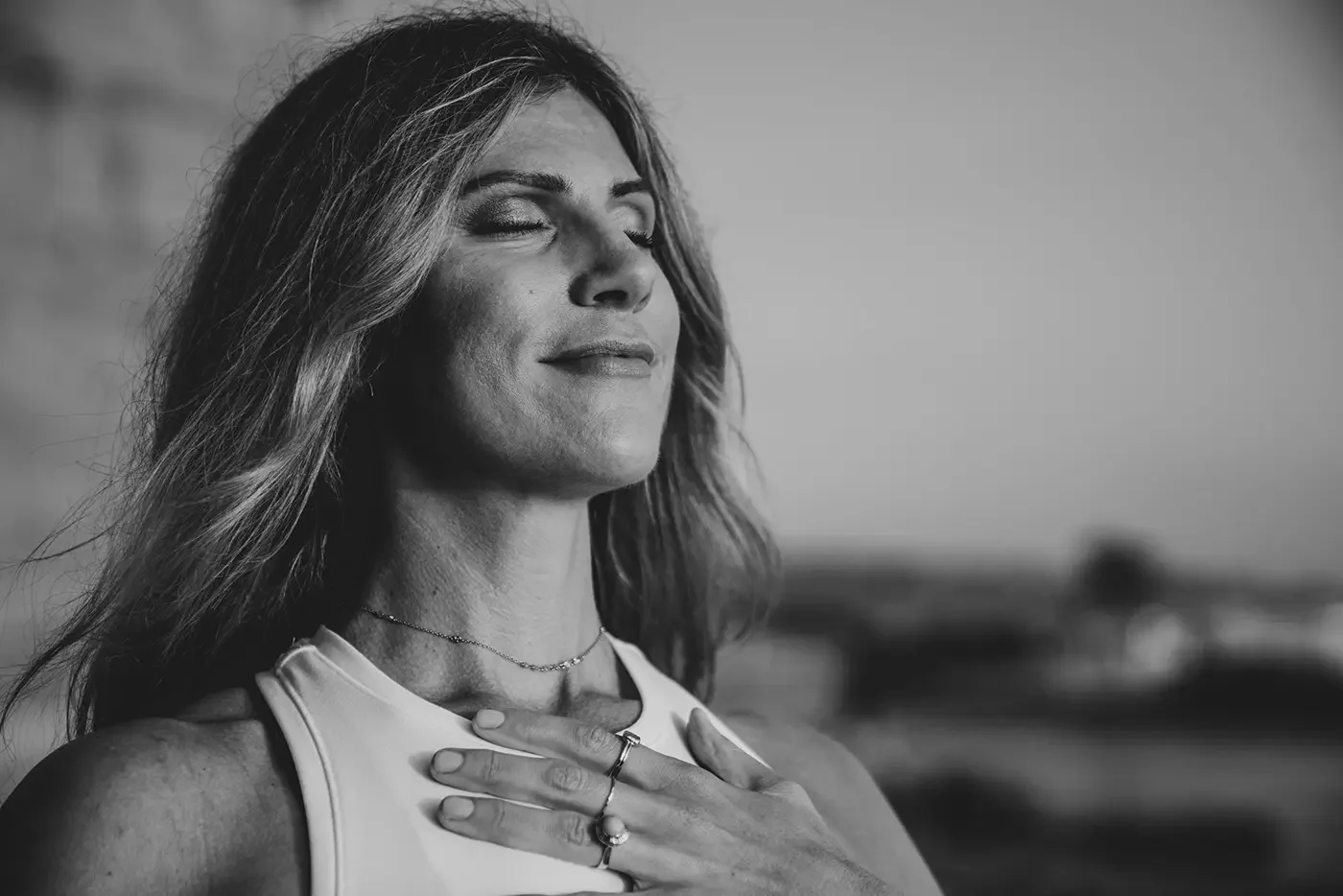 A woman with long hair in a sleeveless top, eyes closed, smiling gently, touching her chest with her right hand, standing outdoors.