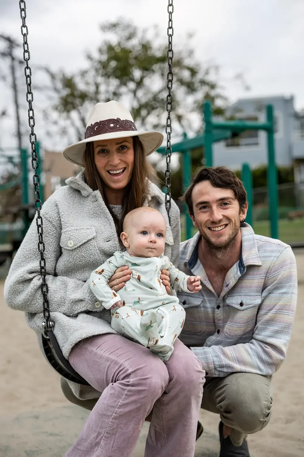 A woman wearing a cream-colored jacket and hat, a man in a plaid shirt, and a baby in pajamas sitting on a swing at a playground, smiling and enjoying the day.