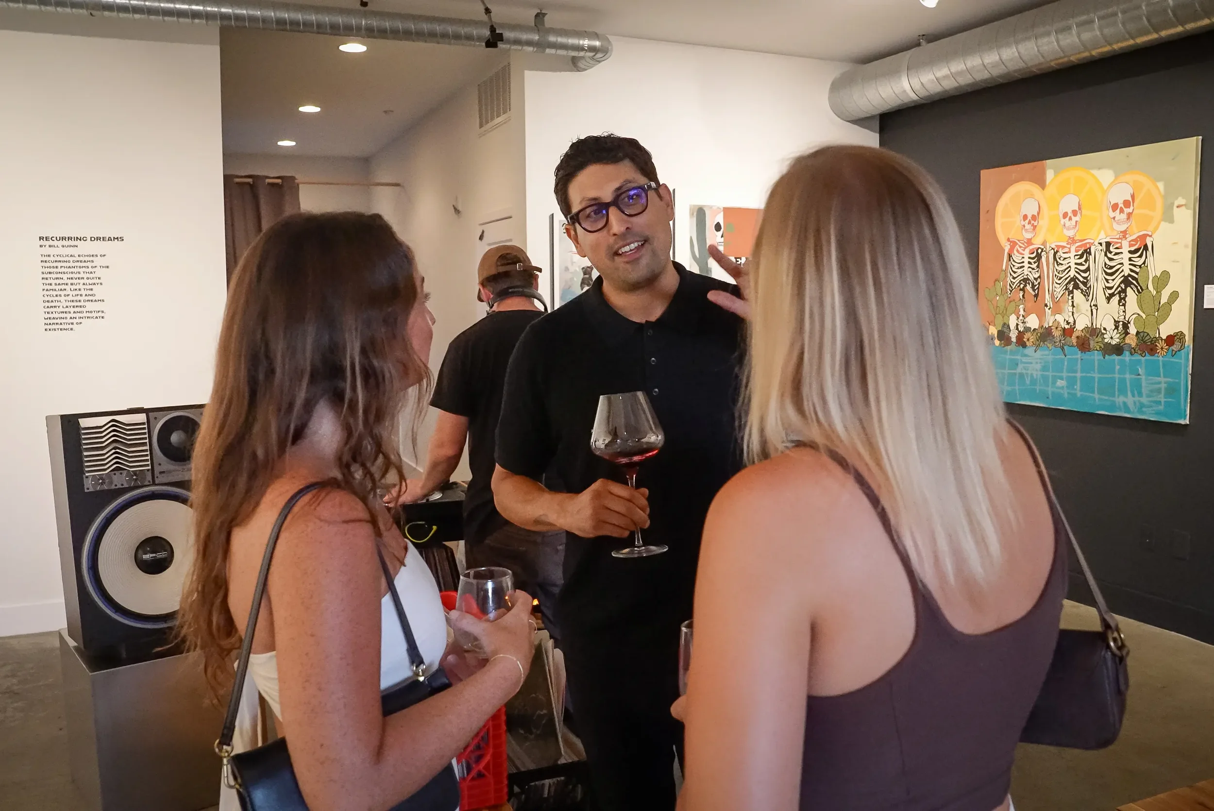 A man in glasses holding a glass of red wine engaging in conversation with three women at an art gallery or exhibition, with artwork and speakers visible in the background.