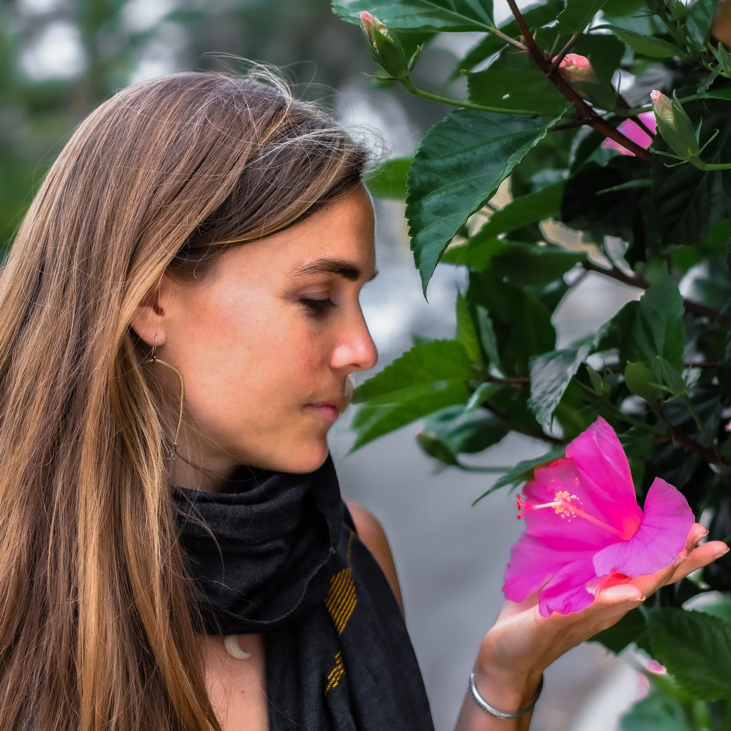 A woman with long light brown hair holding pink hibiscus flowers near her face with green leaves in the background.