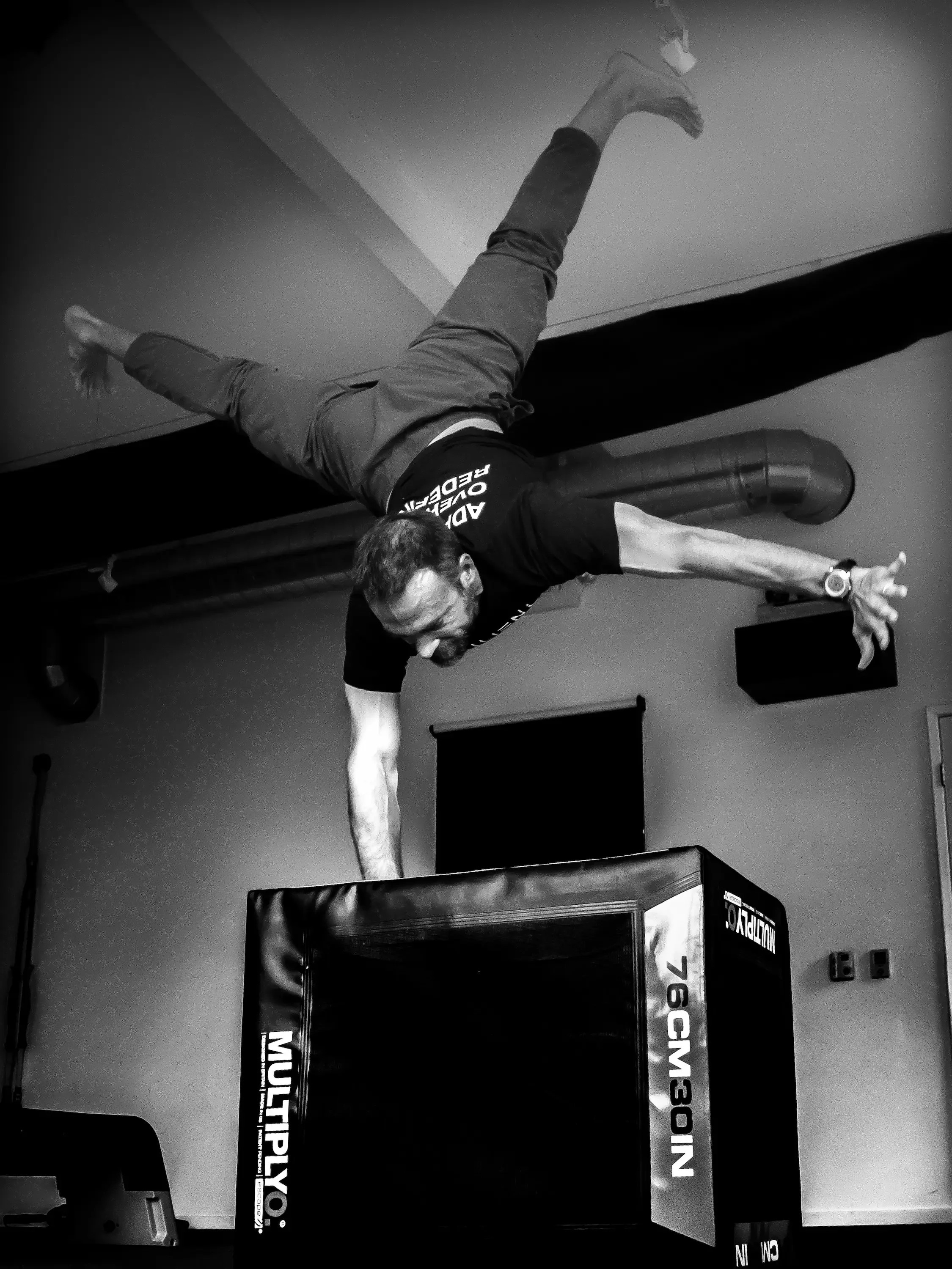 A person performing a handstand on a padded box in an indoor gym or training facility, with the person's body parallel to the ground and legs extended upward.