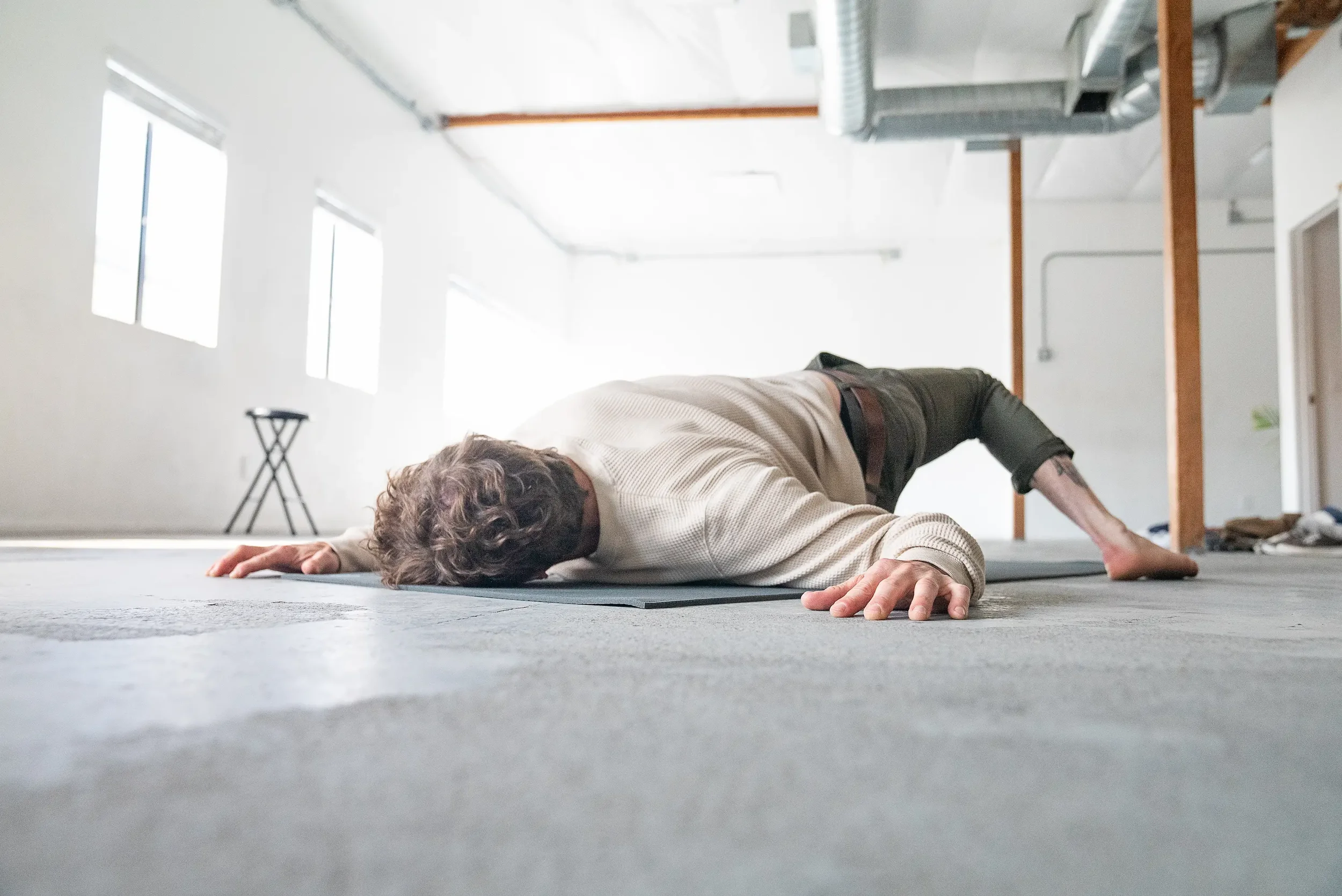 A person practicing yoga or stretching in a bright, spacious room with large windows and industrial-style ductwork.