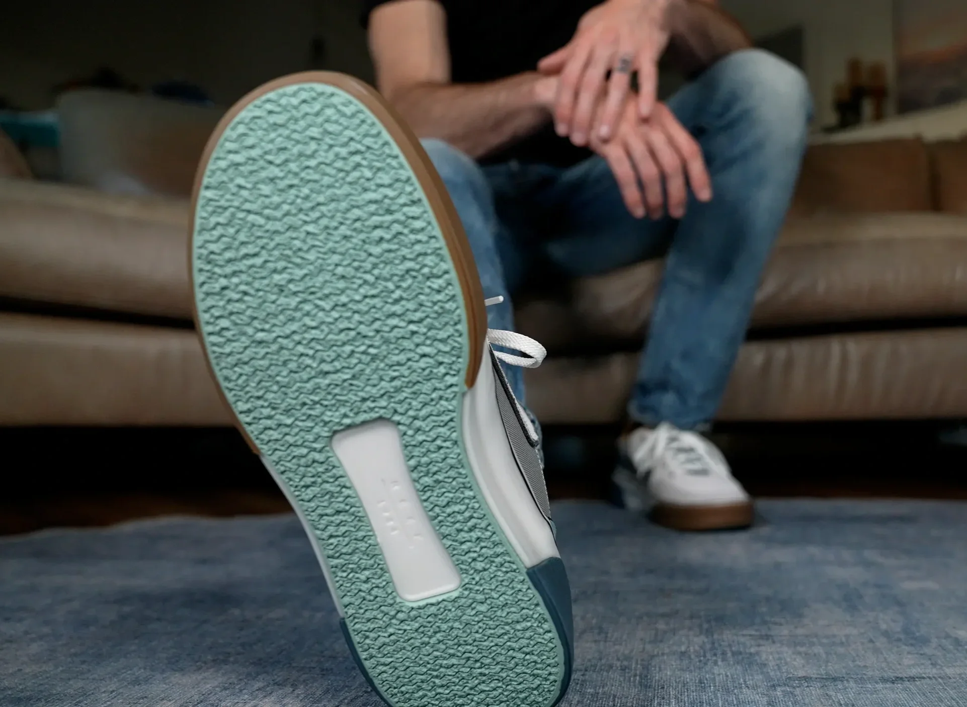 Close-up of a person's foot wearing a white sneaker with a mint green and white sole, resting on a blue carpeted floor, with a person sitting on a couch in the background.