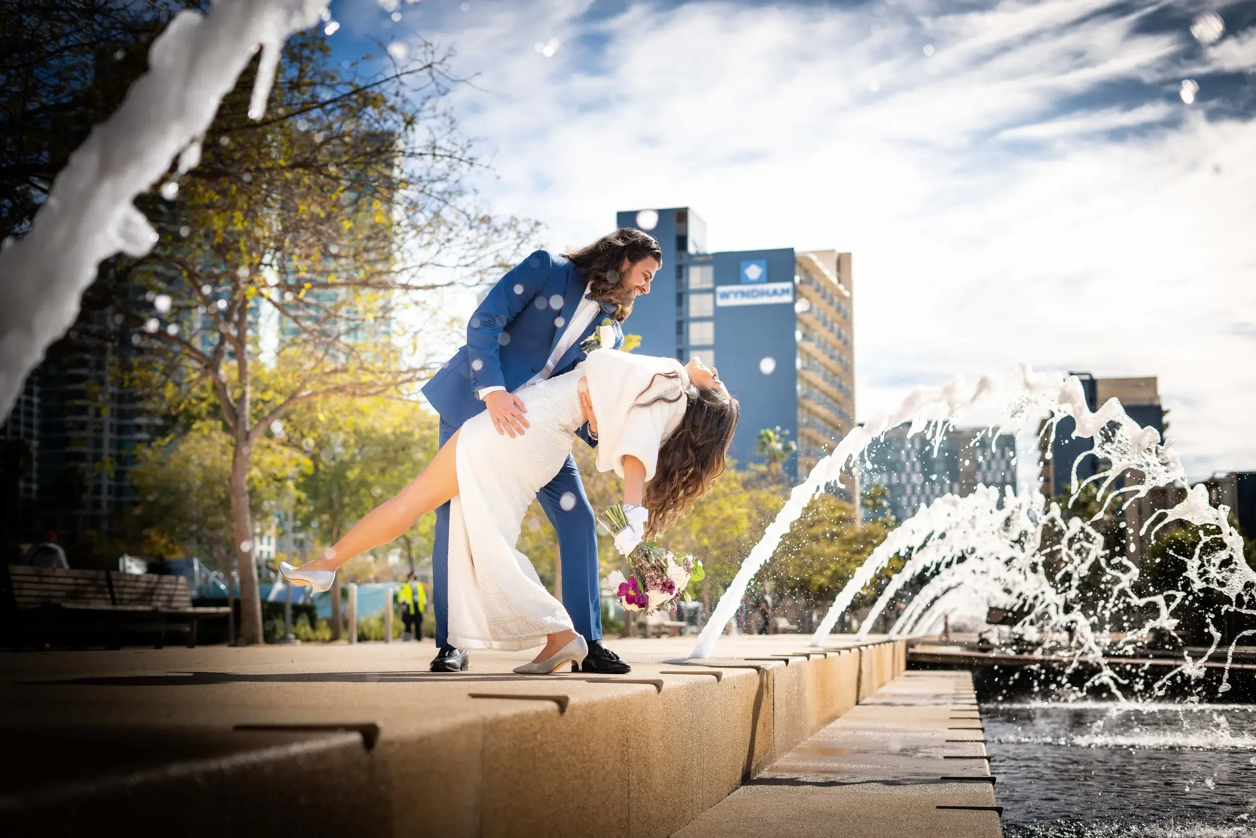 A man in a blue suit and a woman in a white wedding dress dance on a city sidewalk near a fountain, with water splashing around them. The woman holds a bouquet of flowers, and the man supports her as they pose