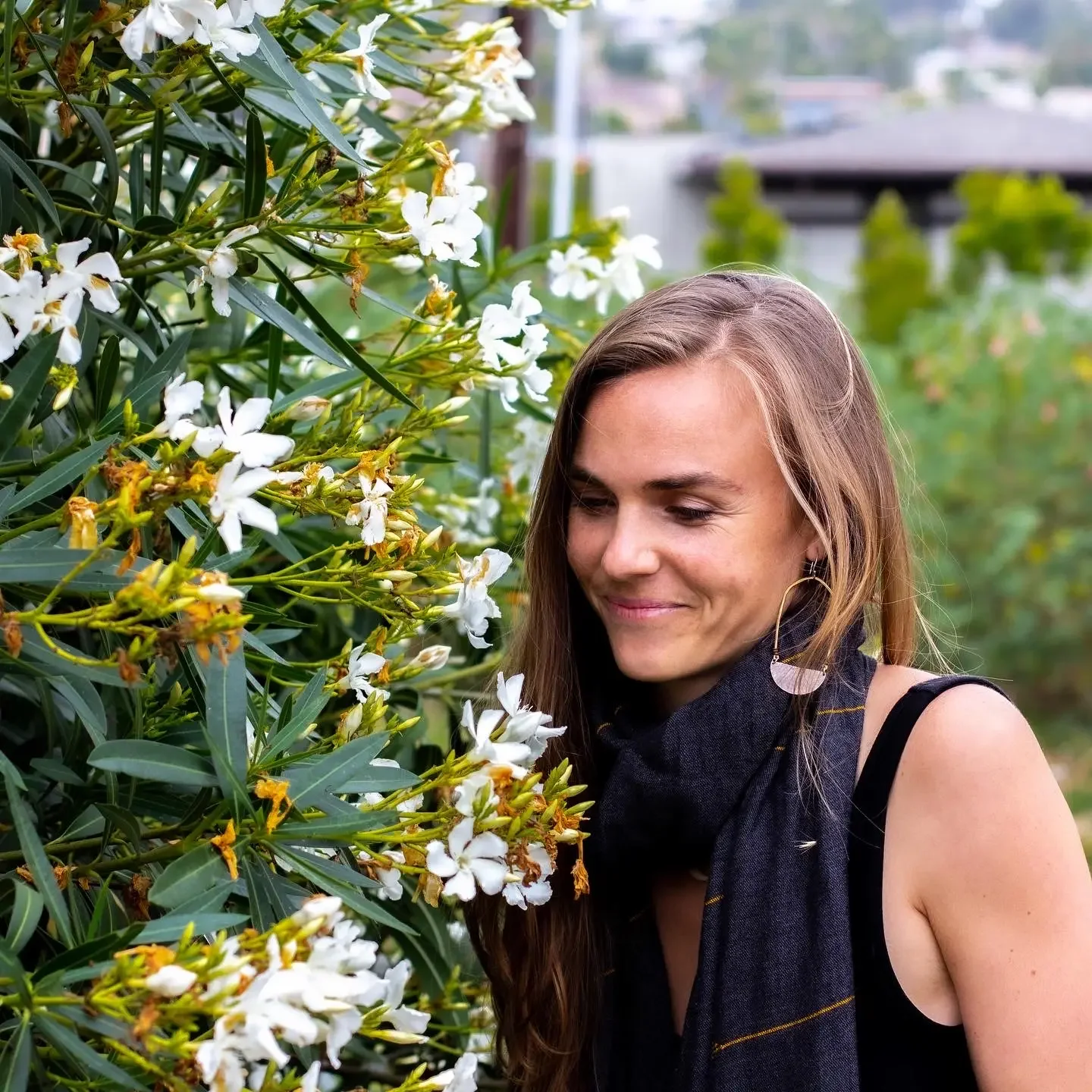 A woman with light brown hair, wearing a black sleeveless top and a black scarf, standing next to a bush with white flowers.