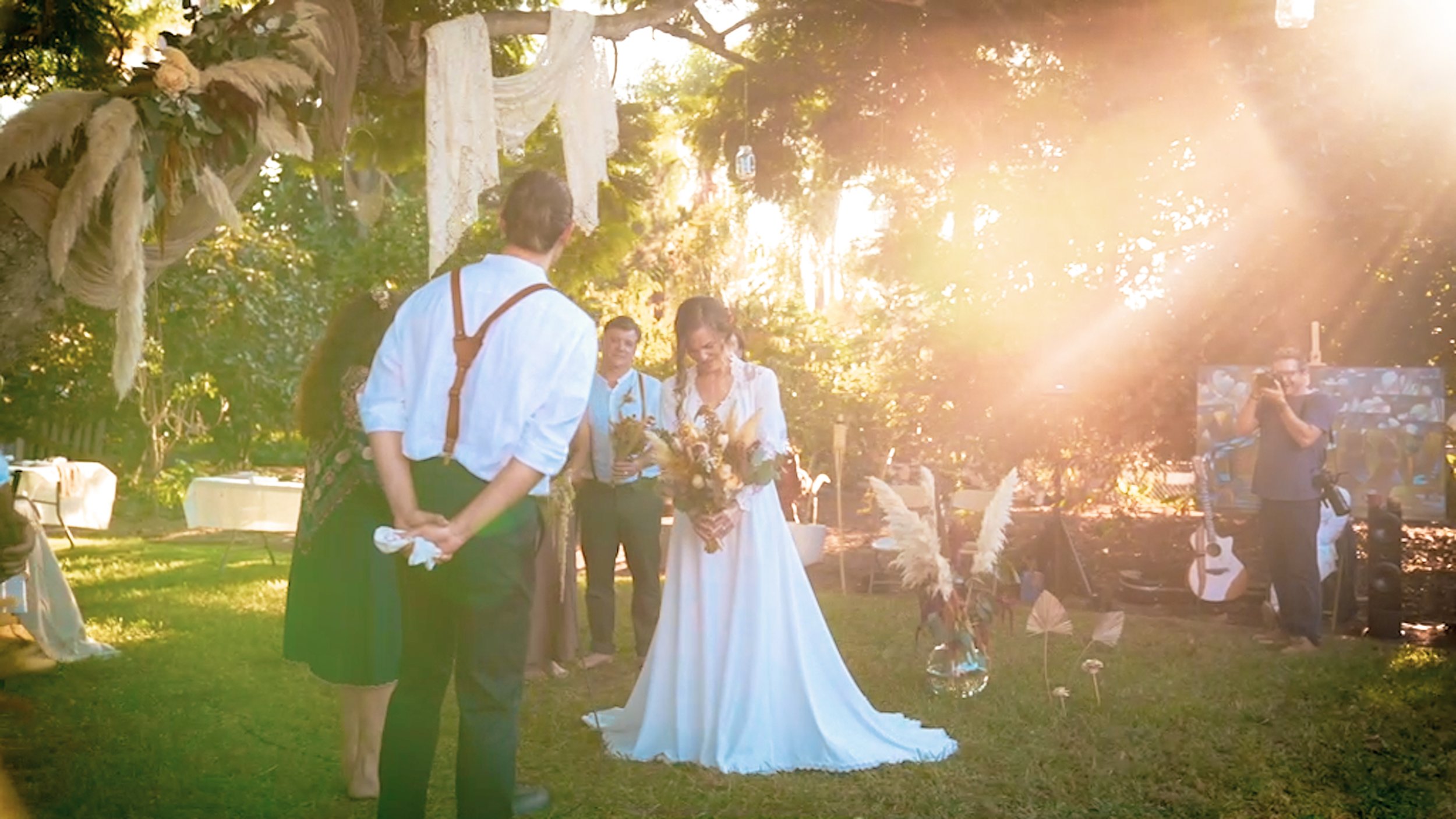 A wedding ceremony outdoors during sunset with people standing around a bride in a white dress holding a bouquet, and a groom in a white shirt. Decor includes white drapery and dried floral arrangements. A guitar and a person taking photos are visible in the background.