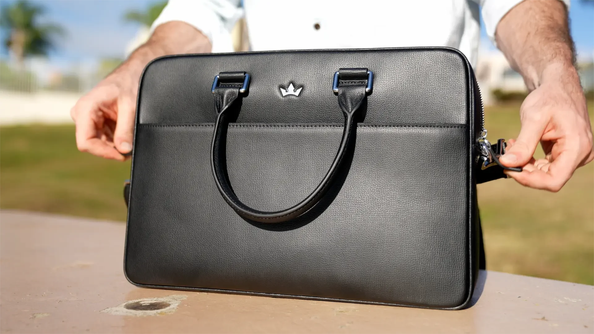Person holding a black leather briefcase outdoors on a sunny day.
