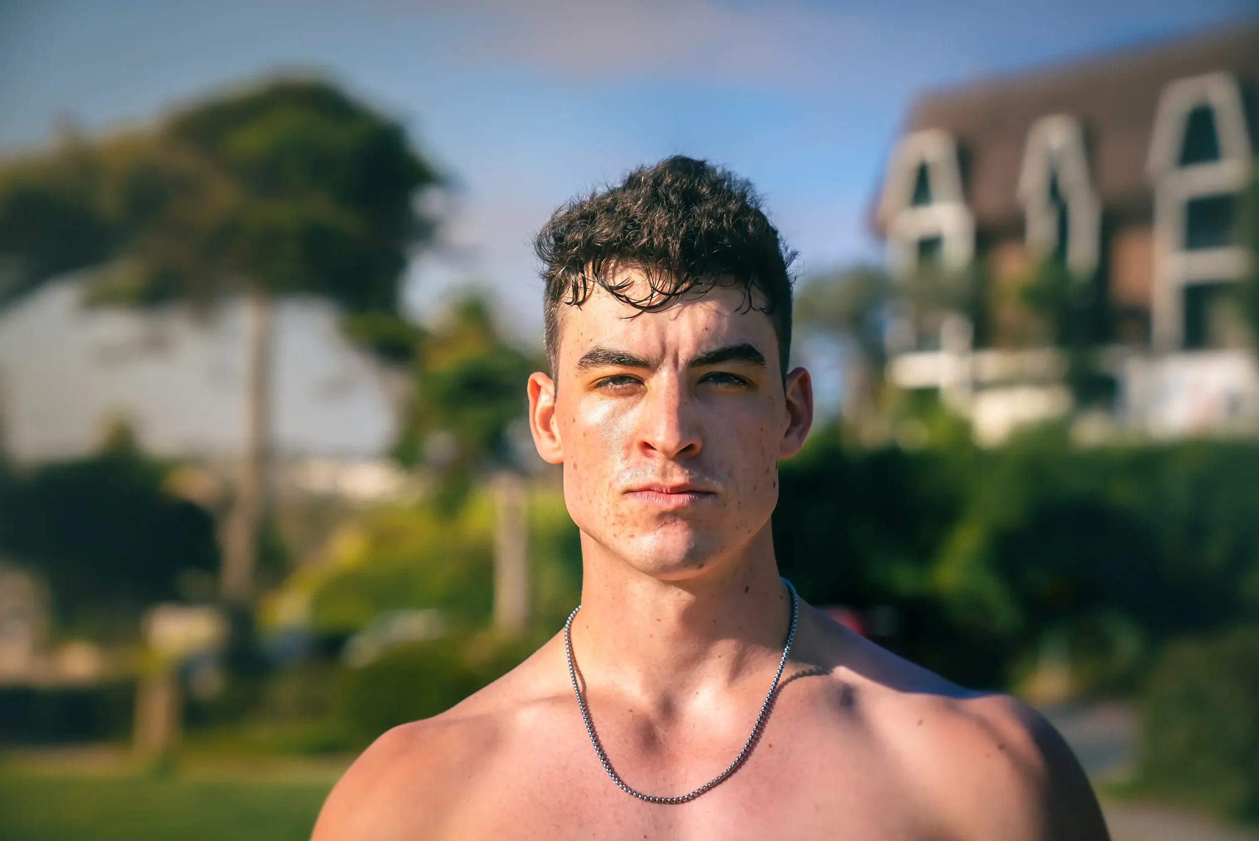 Portrait of a young man with short curly hair, freckles, and a serious expression, outdoors with trees and a house in the background.
