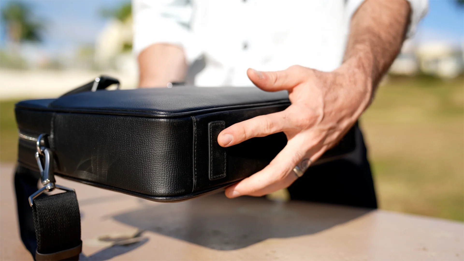 A person's hand holding a black camera bag outdoors with a blurred background of trees and sky.