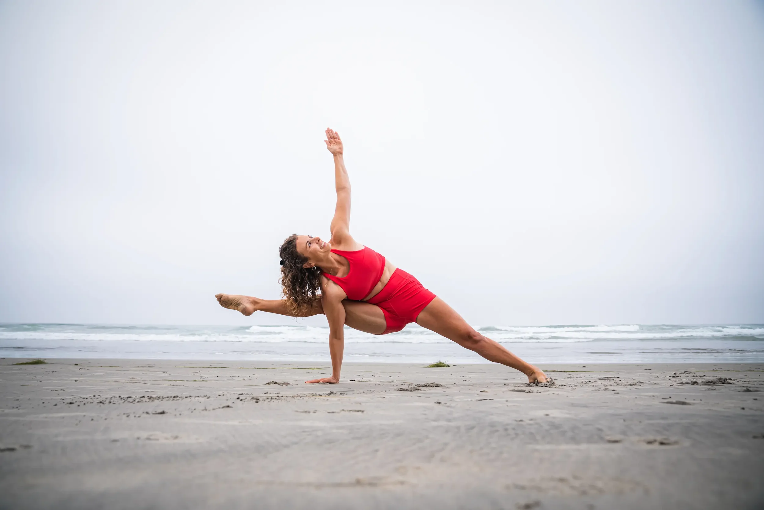 A woman practicing yoga on a beach, balancing in a challenging pose with one hand on the ground, one leg extended behind her, and the other arm reaching upward, wearing a red sports bra and red shorts, with the ocean and cloudy sky in the background.
