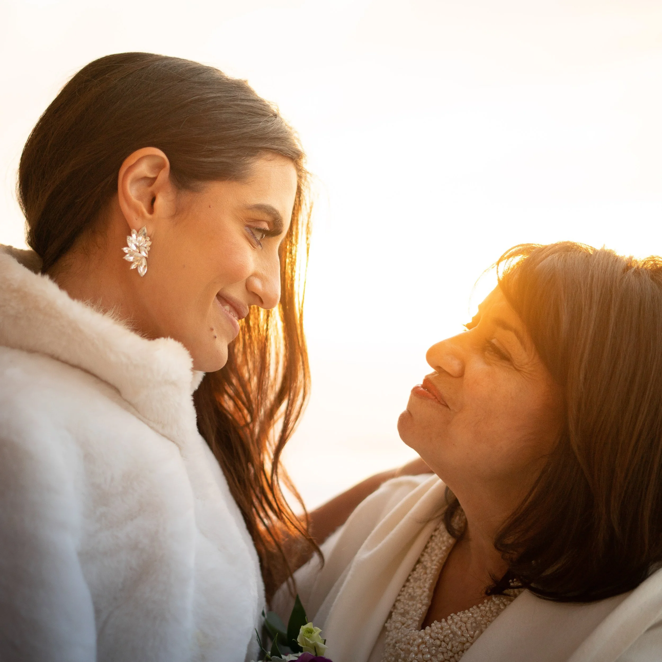 Close-up of a woman with brown hair wearing floral earrings, dressed in a white outfit, and a person with reddish hair in a beige or light-colored outfit, both smiling at each other.
